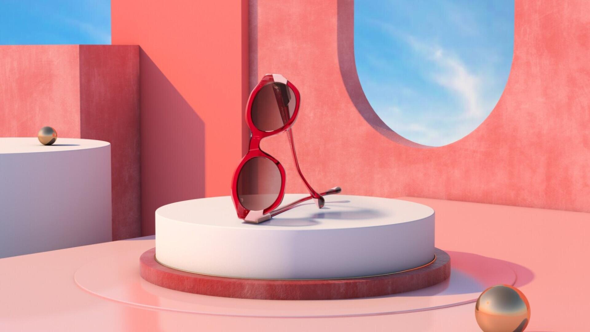 Red sunglasses rest on a white circular pedestal in a minimal, modern pink setting with geometric shapes and a window showing blue sky. Small metallic spheres are placed around the scene.