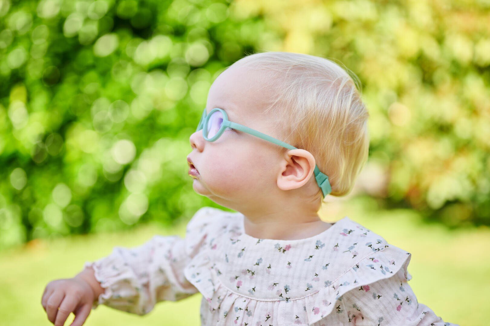 Ein junges Kind mit hellblondem Haar, das eine blaue Brille und ein blumengemustertes Oberteil trägt, ist im Profil im Freien zu sehen, mit einem unscharfen grünen Hintergrund aus Blättern.