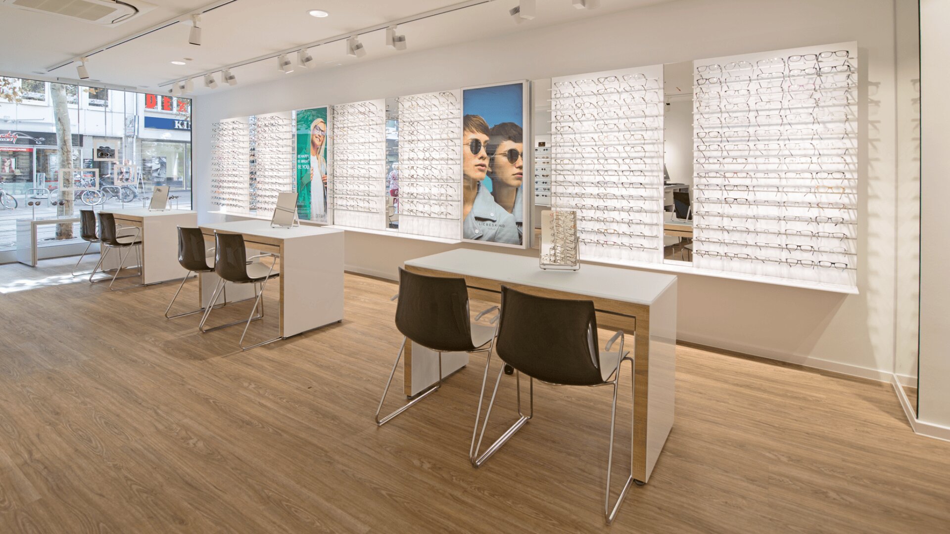 Modernes Optikergeschäft mit großen Ausstellungswänden für Brillen, minimalistischen weißen Schreibtischen mit Stühlen für Kunden, heller Beleuchtung und Holzfußboden. An der Rückwand hängen große Poster von Brillenmodellen.