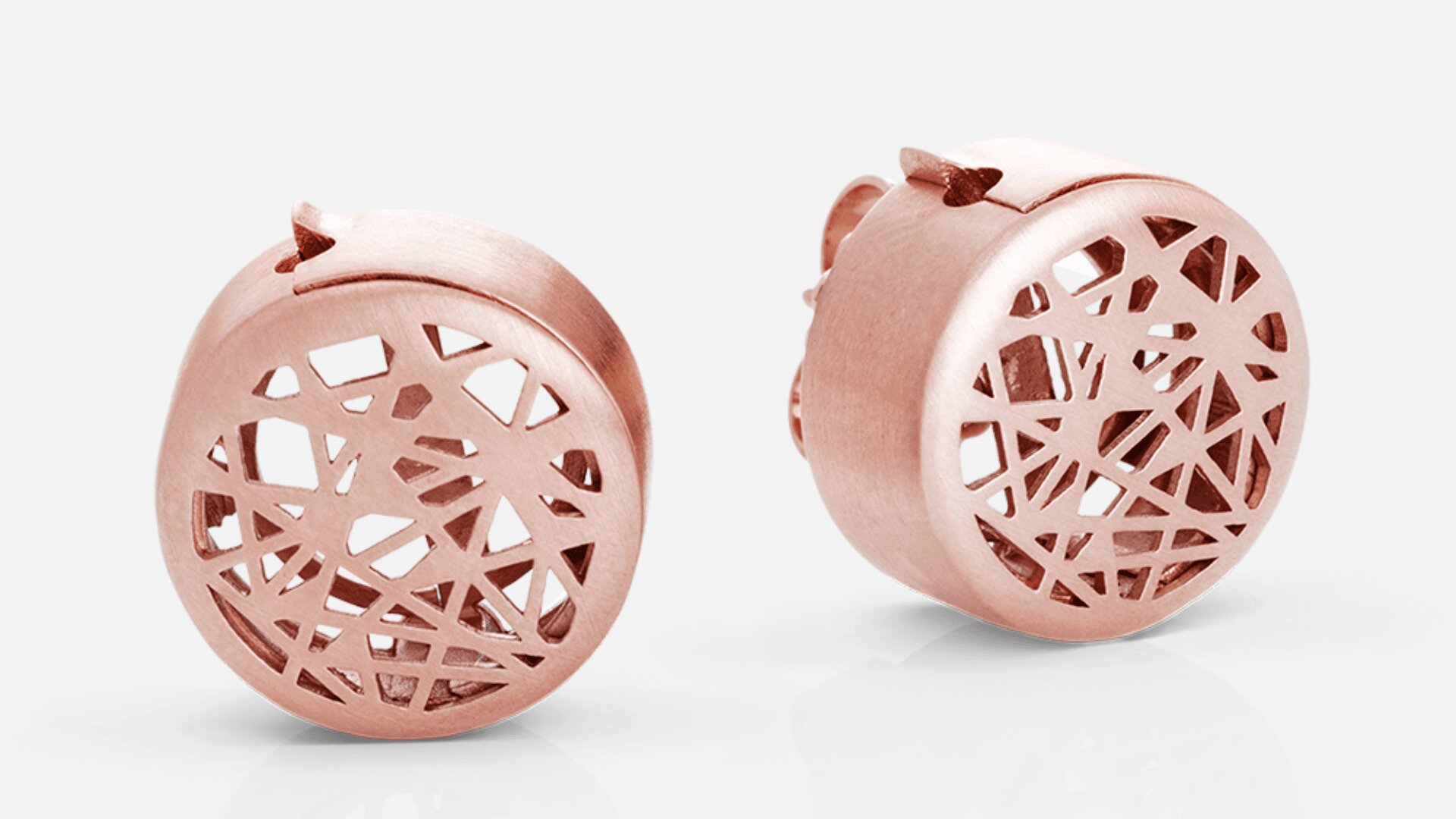 Ein Paar runde Ohrstecker aus Roségold mit einem komplizierten geometrischen Ausschnittmuster auf der Vorderseite, das vor einem schlichten hellen Hintergrund zu sehen ist.