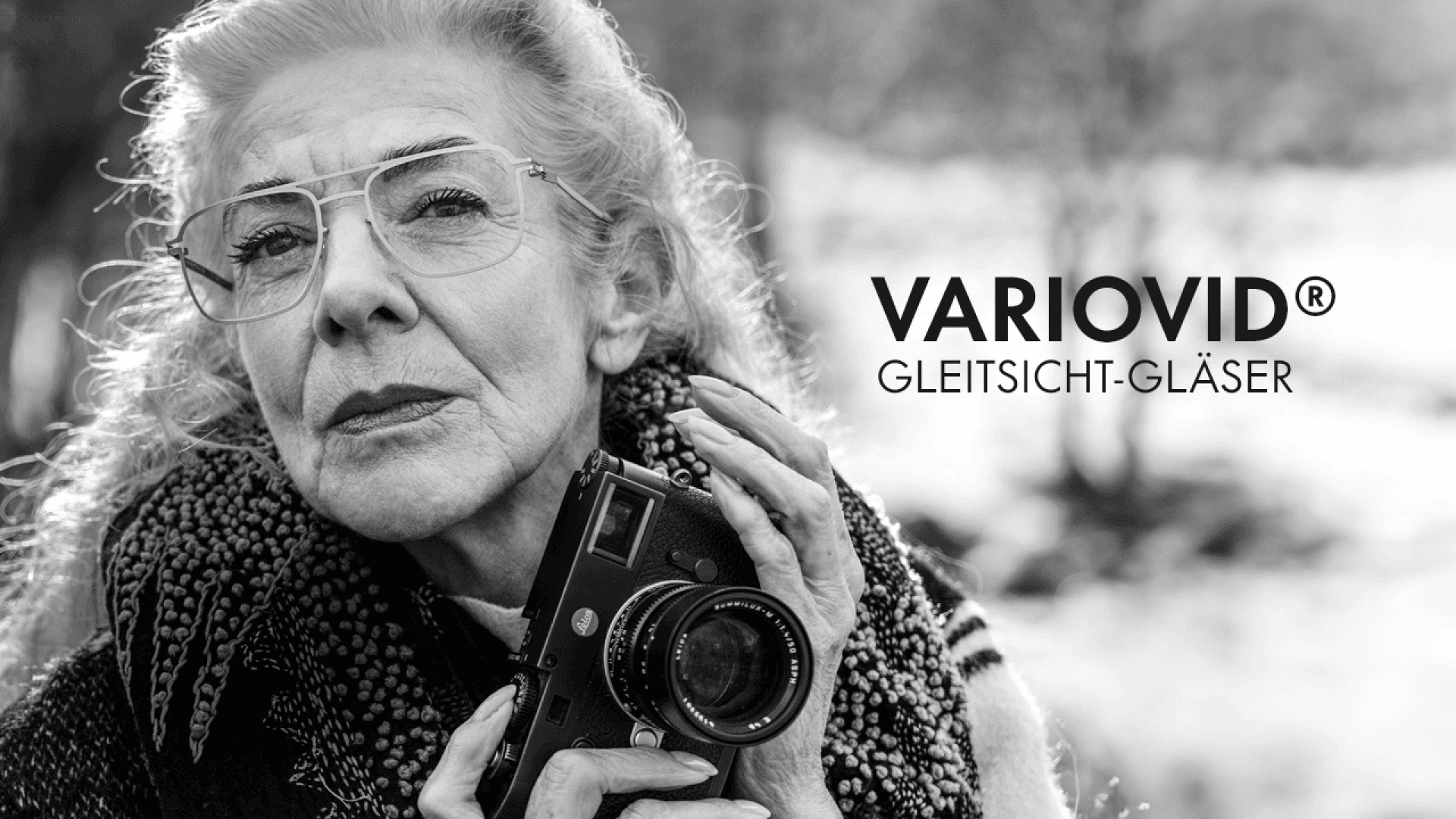 Black and white photo of an elderly woman wearing glasses and holding a camera, looking thoughtfully into the distance. Text reads: VARIOVID® Gleitsicht-Gläser.