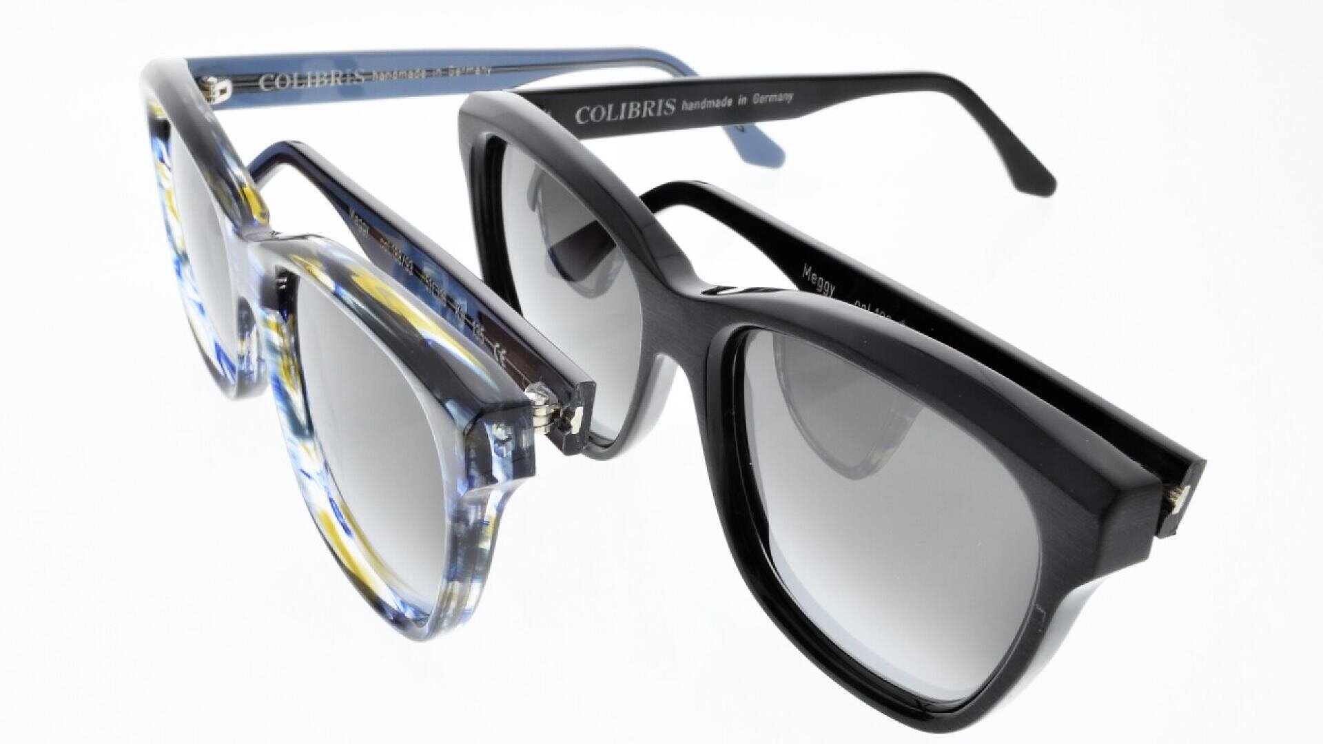 Two pairs of eyeglasses with thick frames, one in a translucent blue and yellow pattern and the other in solid black, both with the brand name “COLIBRIS” visible on the arms, displayed on a white background.