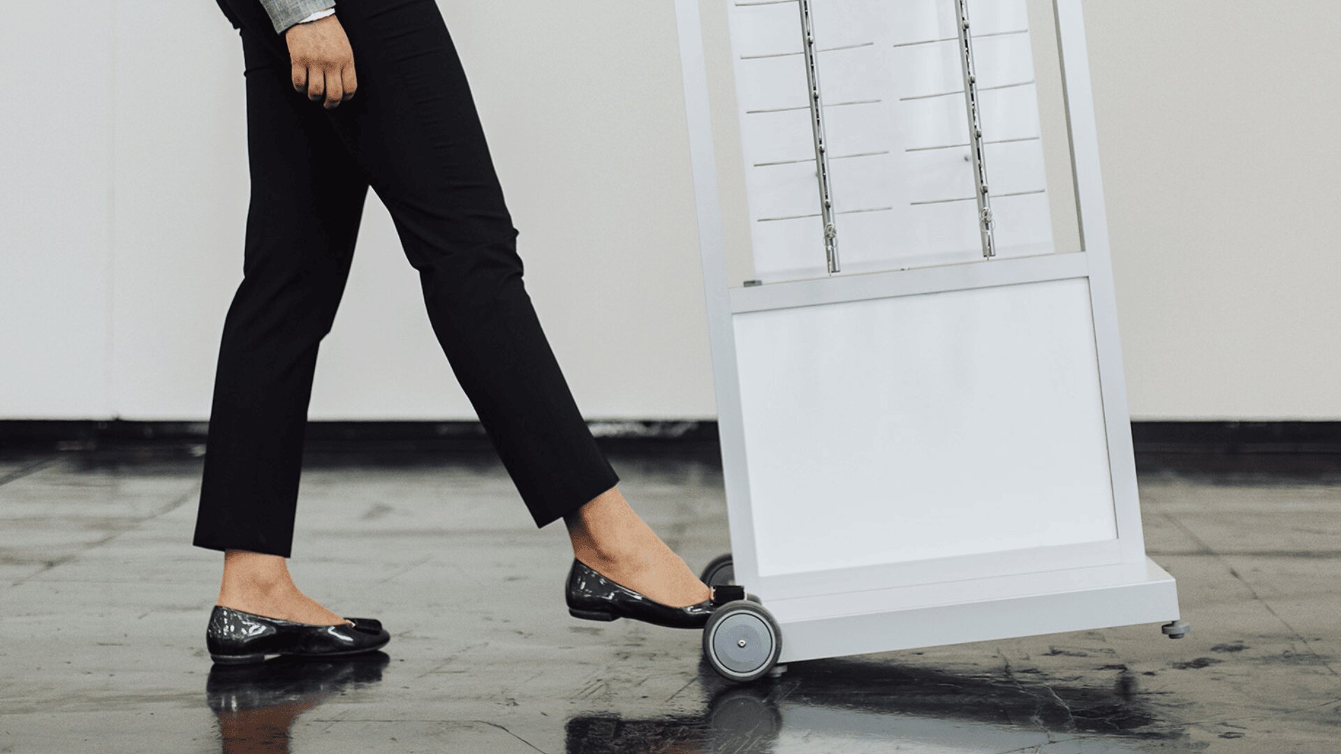 A person wearing black pants and black flats uses their foot to tilt a white cart with wheels on a shiny floor.