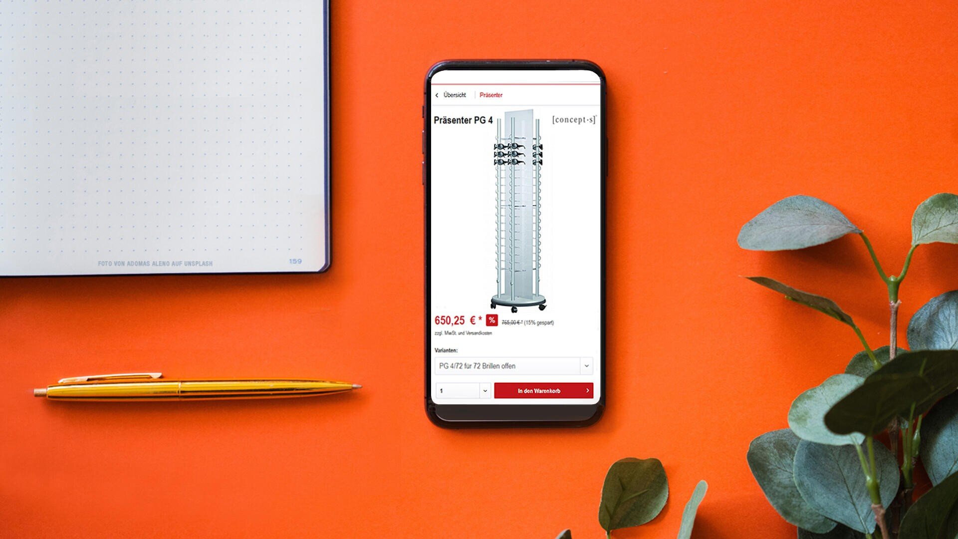 Ein Smartphone zeigt eine Produktseite für einen drehbaren Displayständer auf einem orangefarbenen Schreibtisch an, daneben liegen ein Notizbuch, zwei gelbe Bleistifte und einige grüne Blätter.