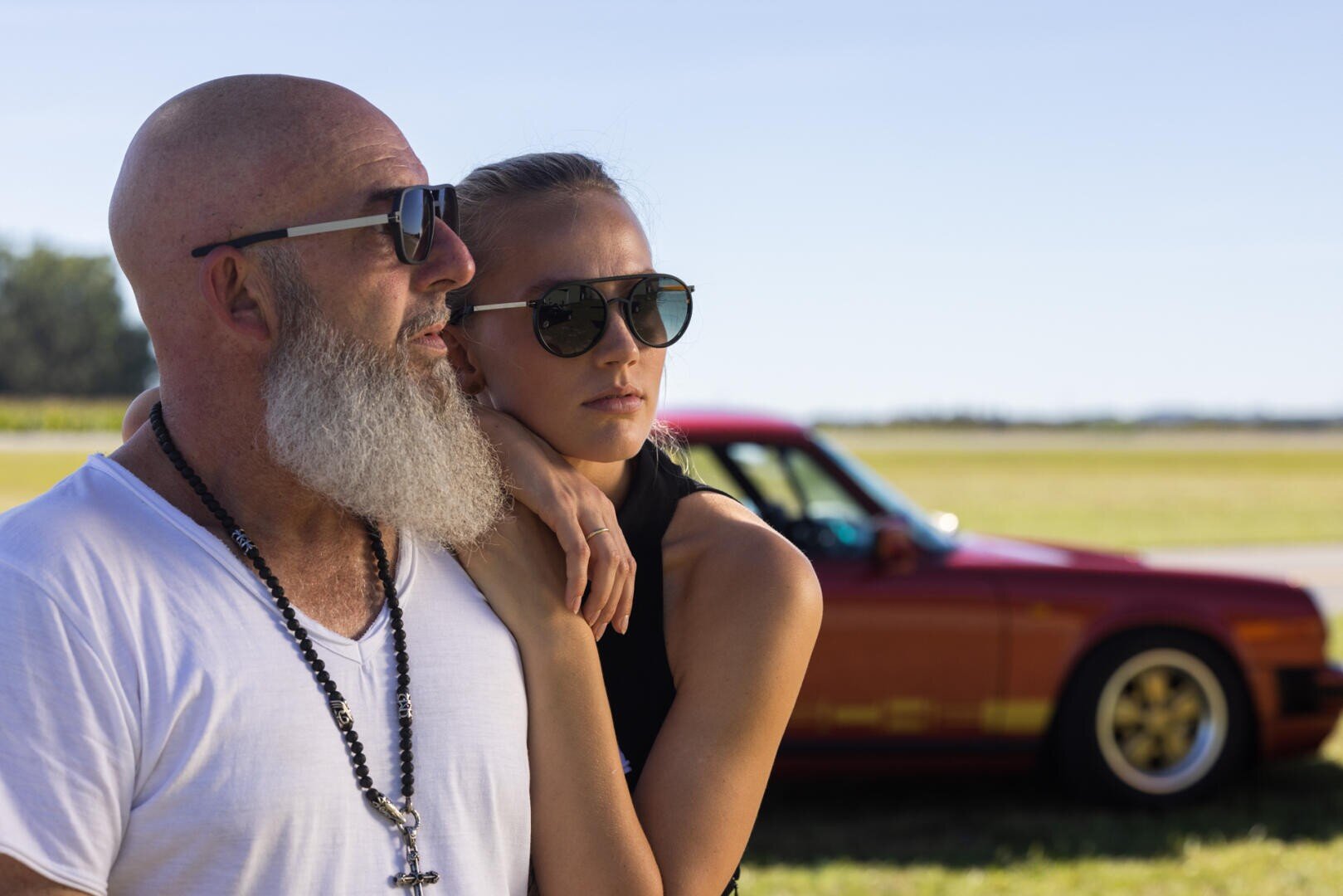 Ein bärtiger Mann mit Sonnenbrille und weißem Hemd mit einer Kreuzkette steht dicht neben einer Frau mit schwarzer Sonnenbrille; im Hintergrund parkt ein rotes Auto an einem sonnigen Tag.