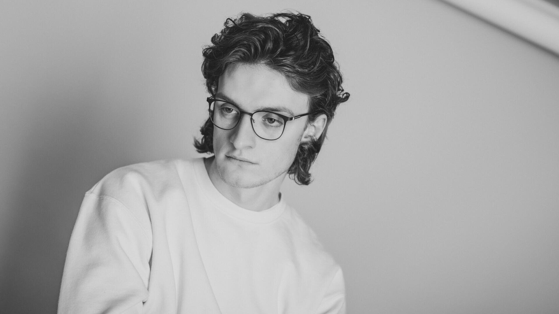 Schwarzweißfoto einer jungen Person mit welligem Haar, Brille und schlichtem Sweatshirt, die nachdenklich vor einem schlichten Hintergrund nach unten blickt.