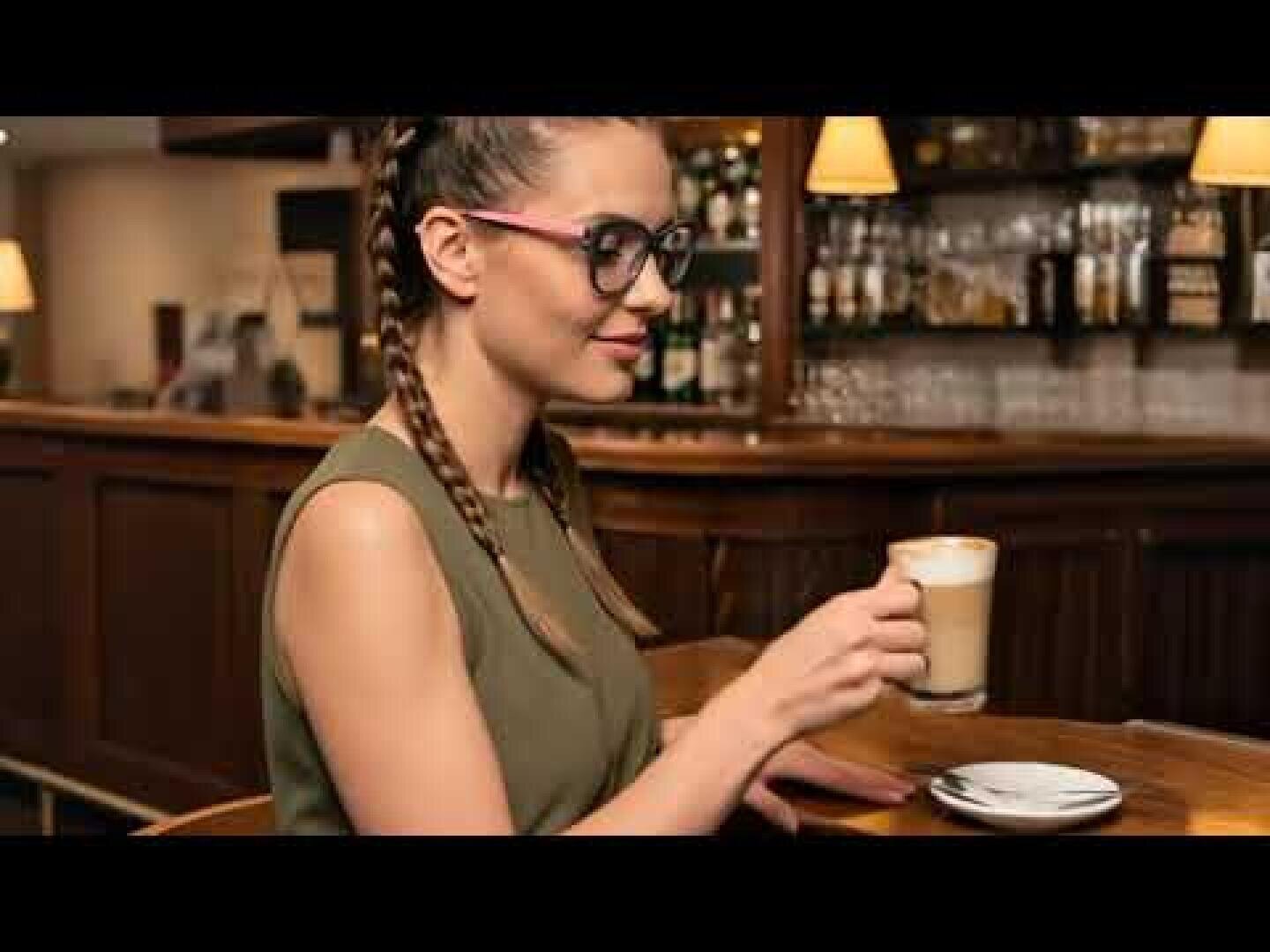 A woman with braided hair and glasses sits at a wooden bar, a cup of coffee or latte in her hand, shelves with bottles and warm light in the background.