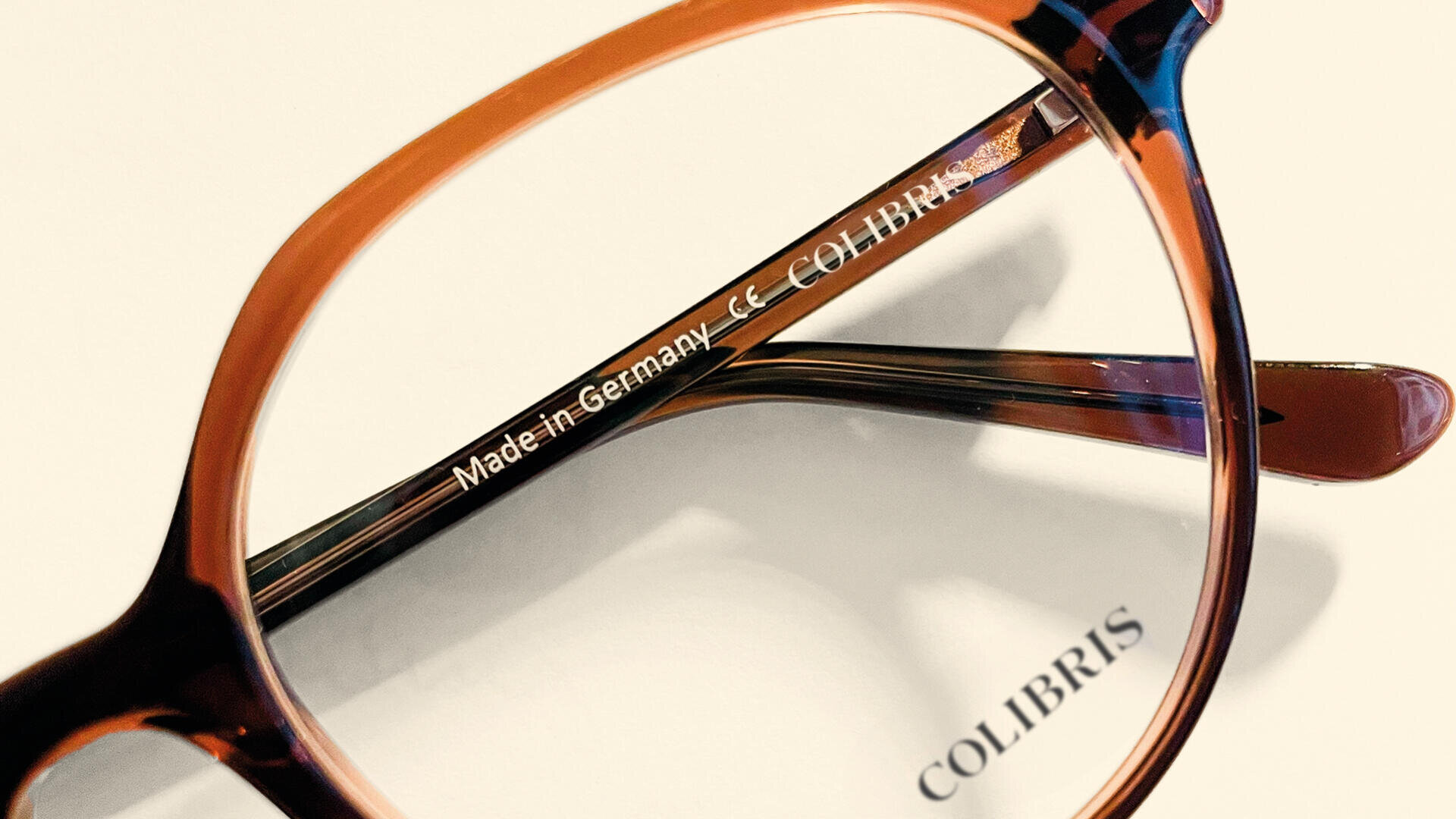 Nahaufnahme einer Brille aus braunem Schildpatt mit dem Text „Made in Germany CE COLIBRIS“ auf der Innenseite eines Bügels und dem Aufdruck „COLIBRIS“ auf der weißen Oberfläche unter den Gläsern.