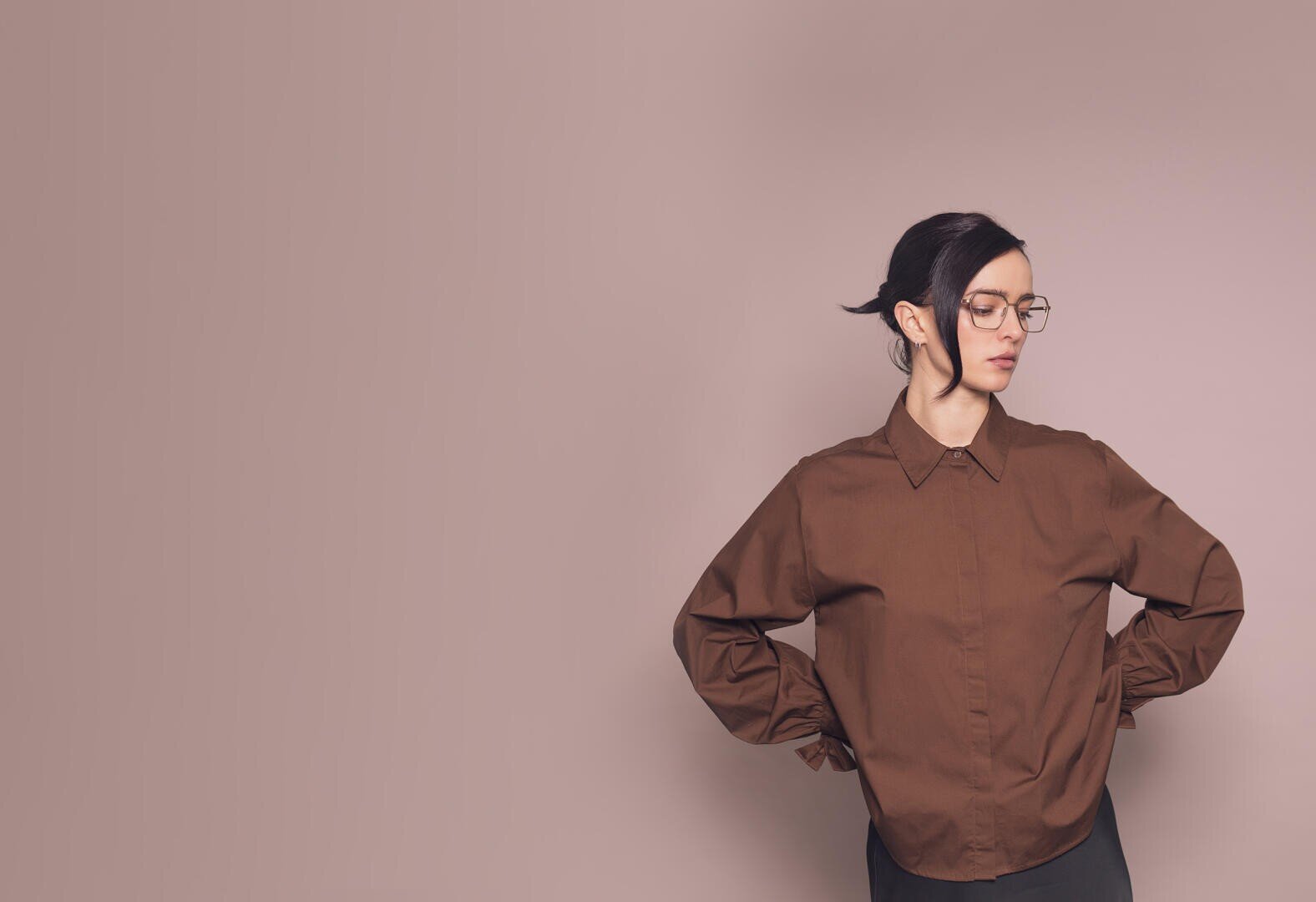 A person with glasses and dark hair, wearing a brown shirt, stands in front of a plain light brown background, hands on hips and looking to the side.