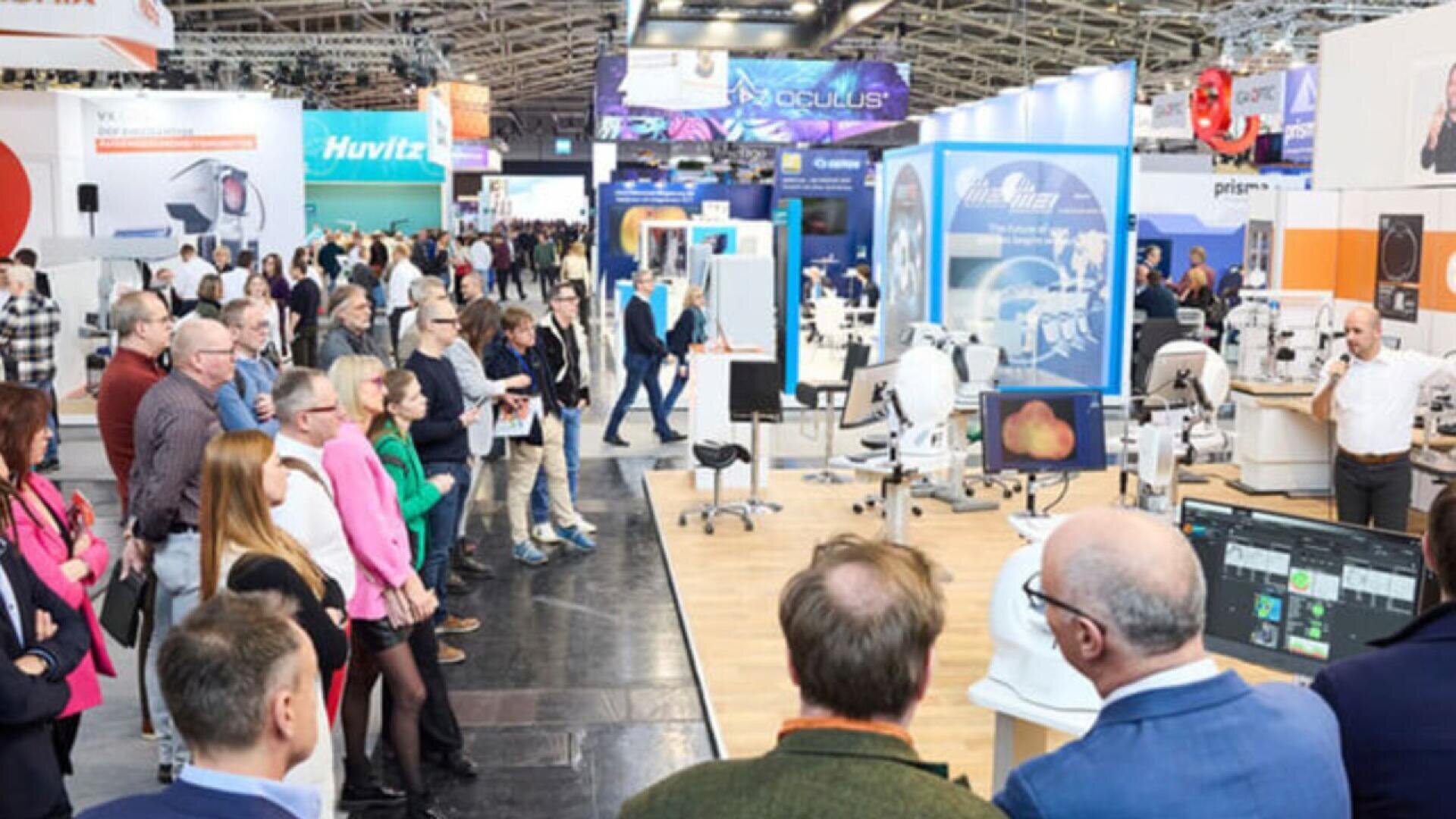 A crowd of people stands in an exhibition hall, attentively watching a presentation on medical or optical equipment, with various booths and technology displays visible in the background.