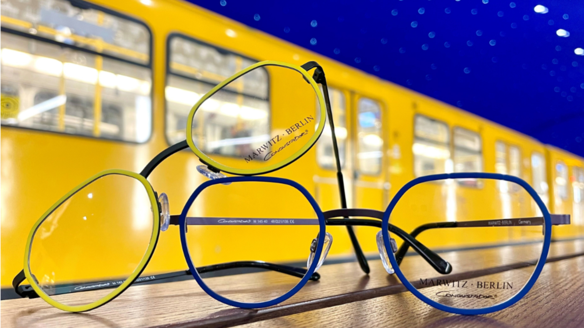 Two pairs of colorful eyeglasses, one green and one blue, rest on a wooden surface with a bright yellow subway train and starry blue ceiling in the background. The eyeglasses show the brand Mawitiz Berlin.