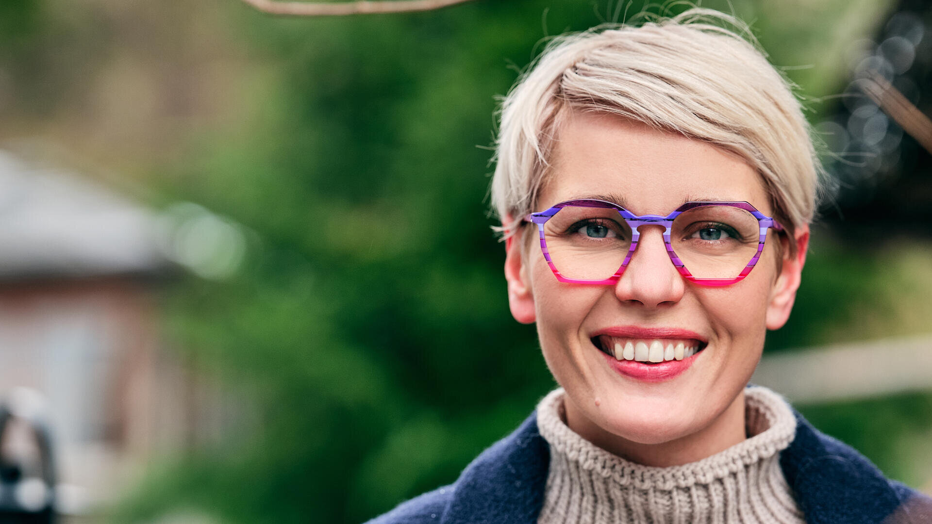 Lächelnde Person mit kurzen blonden Haaren, einer markanten rot-violetten Brille, einem hellbraunen Rollkragenpullover und einem dunklen Mantel, die im Freien steht, mit viel Grün im unscharfen Hintergrund.