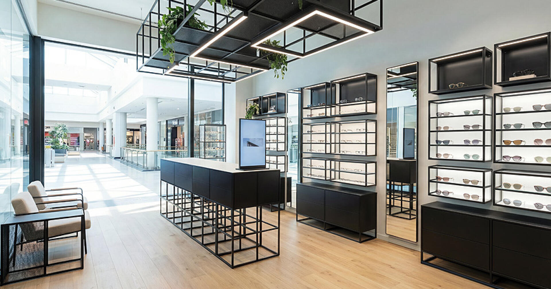 A modern, minimalist eyewear store with black shelves of glasses, large mirrors, an elegant counter and natural light coming in through the floor-to-ceiling windows. There are two chairs in the waiting area.