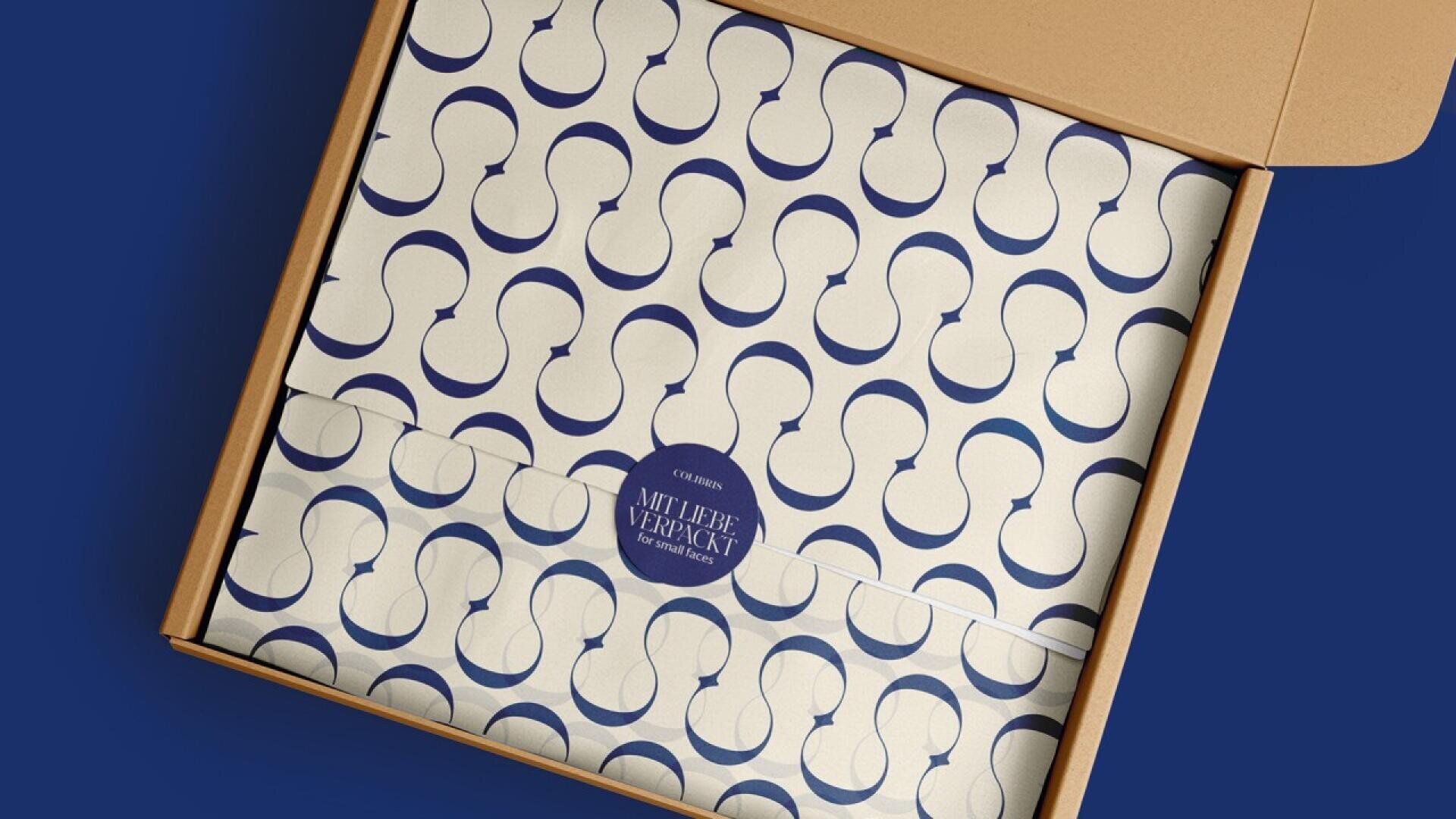 An open cardboard box reveals tissue paper with a blue abstract pattern and a round blue sticker labeled “MULTIPLE VERPACKT” securing the paper.