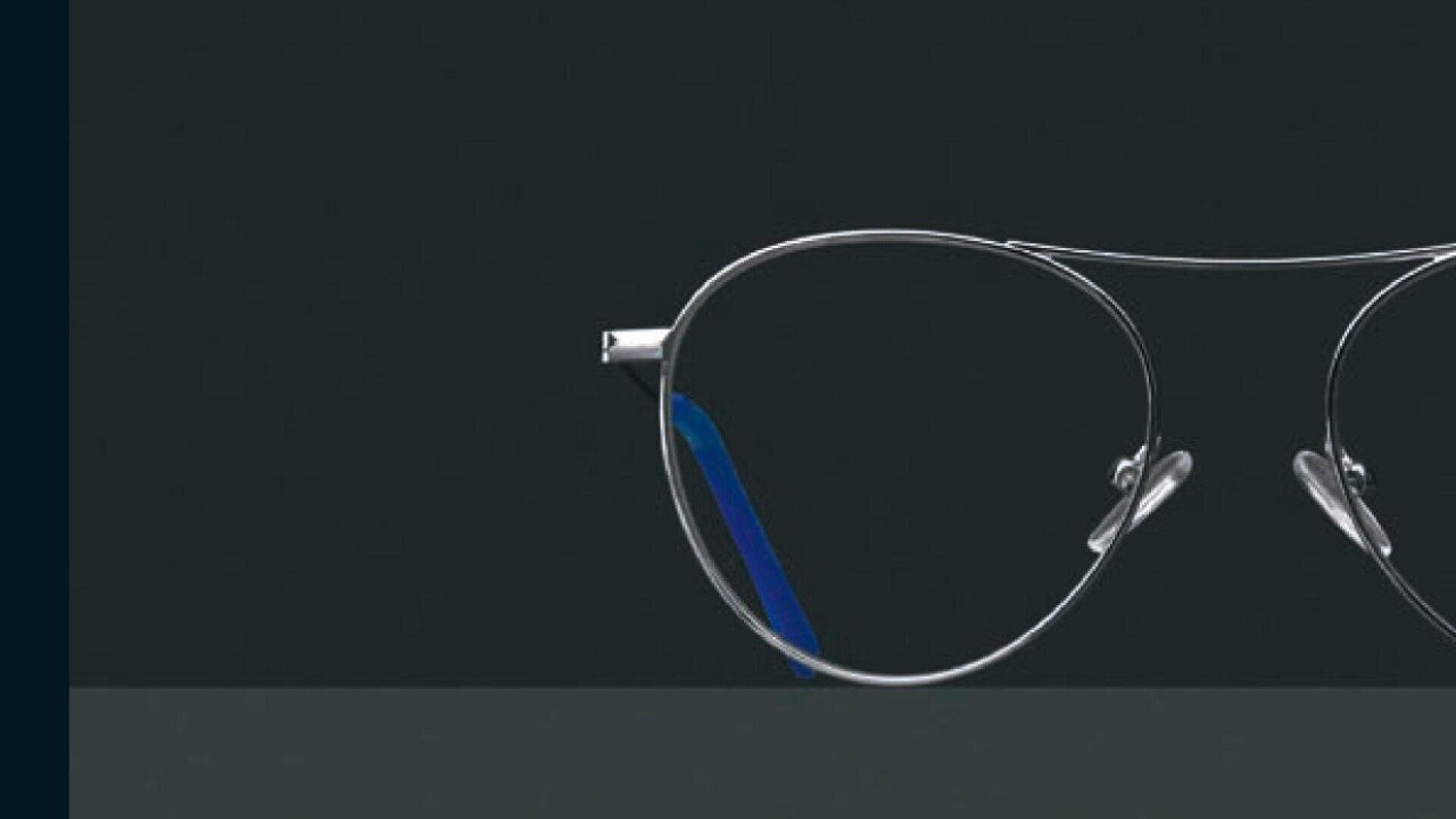 A close-up of the right half of a pair of silver wireframe eyeglasses with blue accents, set against a dark background.