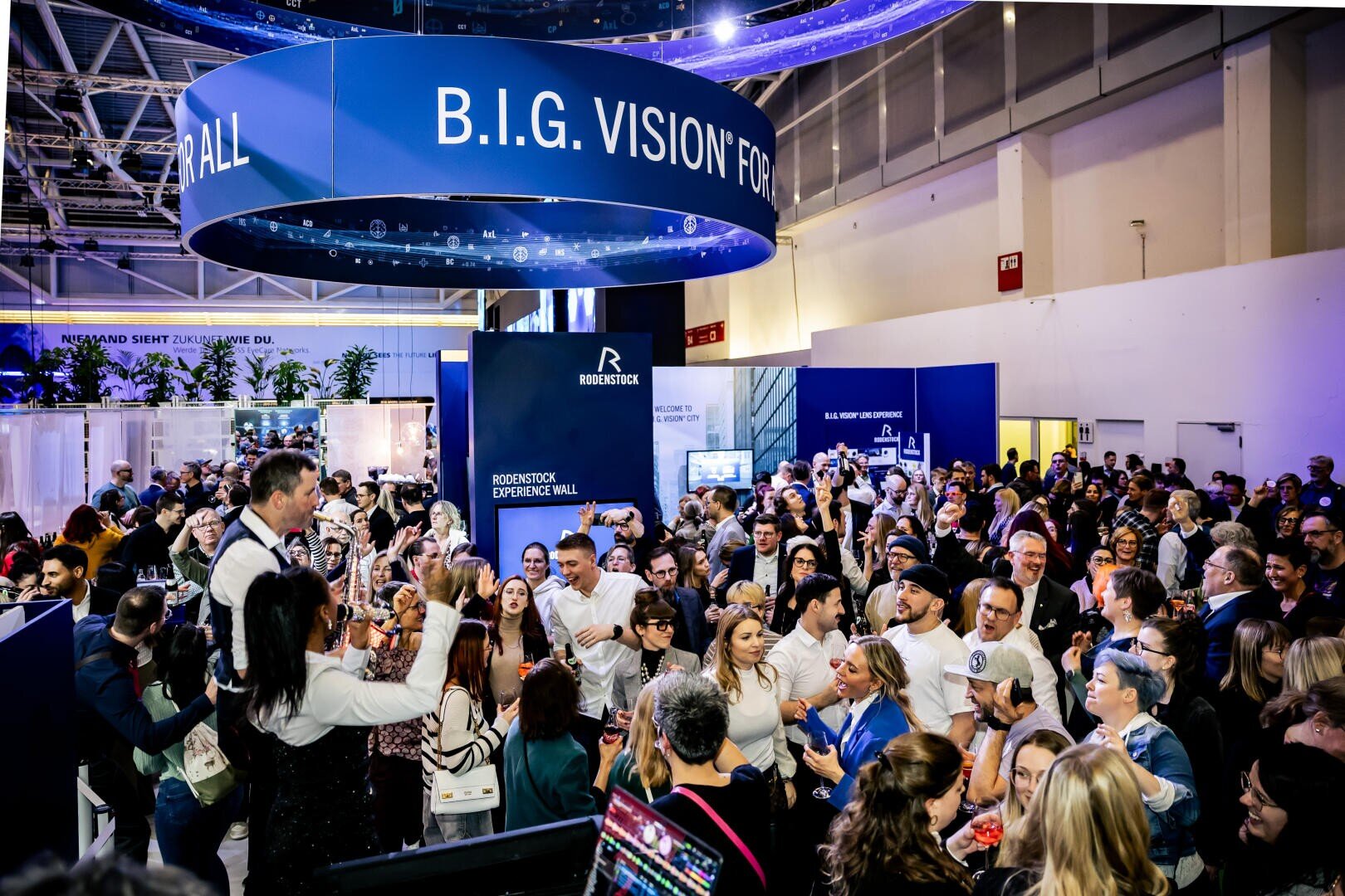 Eine große Menschenmenge versammelt sich auf einer Indoor-Messe unter einem runden Schild mit der Aufschrift B.I.G. VISION. Die Menschen mischen sich, unterhalten sich und machen Fotos vor den modernen Ständen und Auslagen.