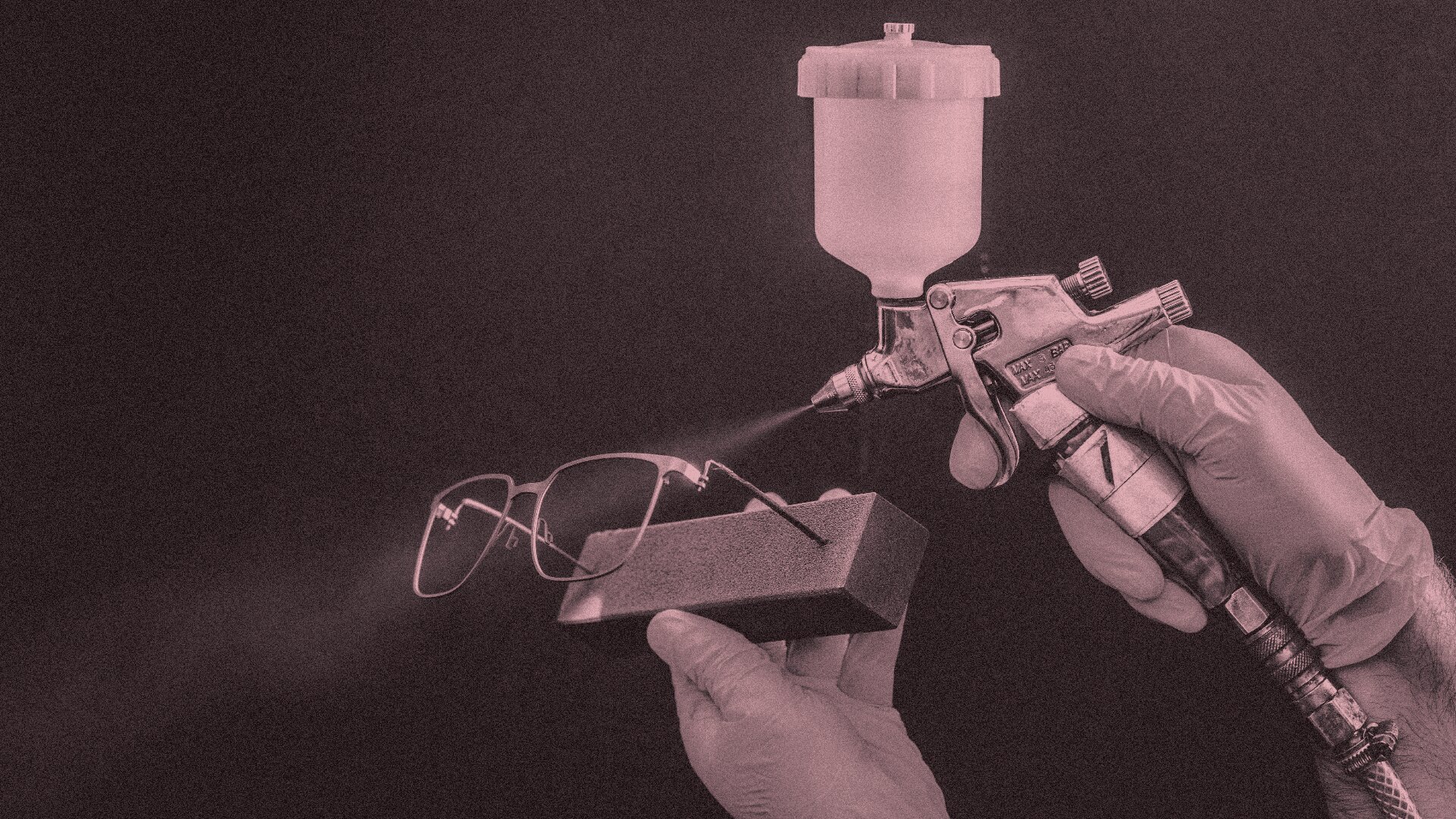 Eine Person mit Handschuhen beschichtet mit einer Spritzpistole eine Brille, die aufrecht auf einem kleinen Ständer vor einem dunklen Hintergrund steht.