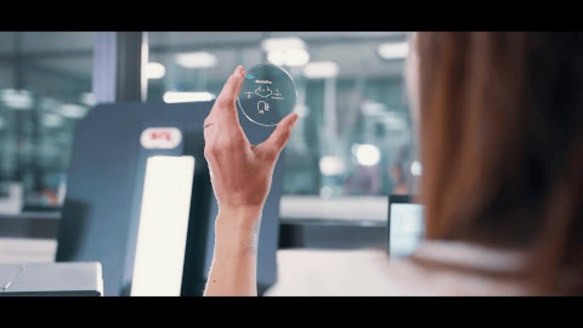 In a bright, modern laboratory or production environment, a person holds up a transparent, round lens or glass with small, white, printed technical symbols.