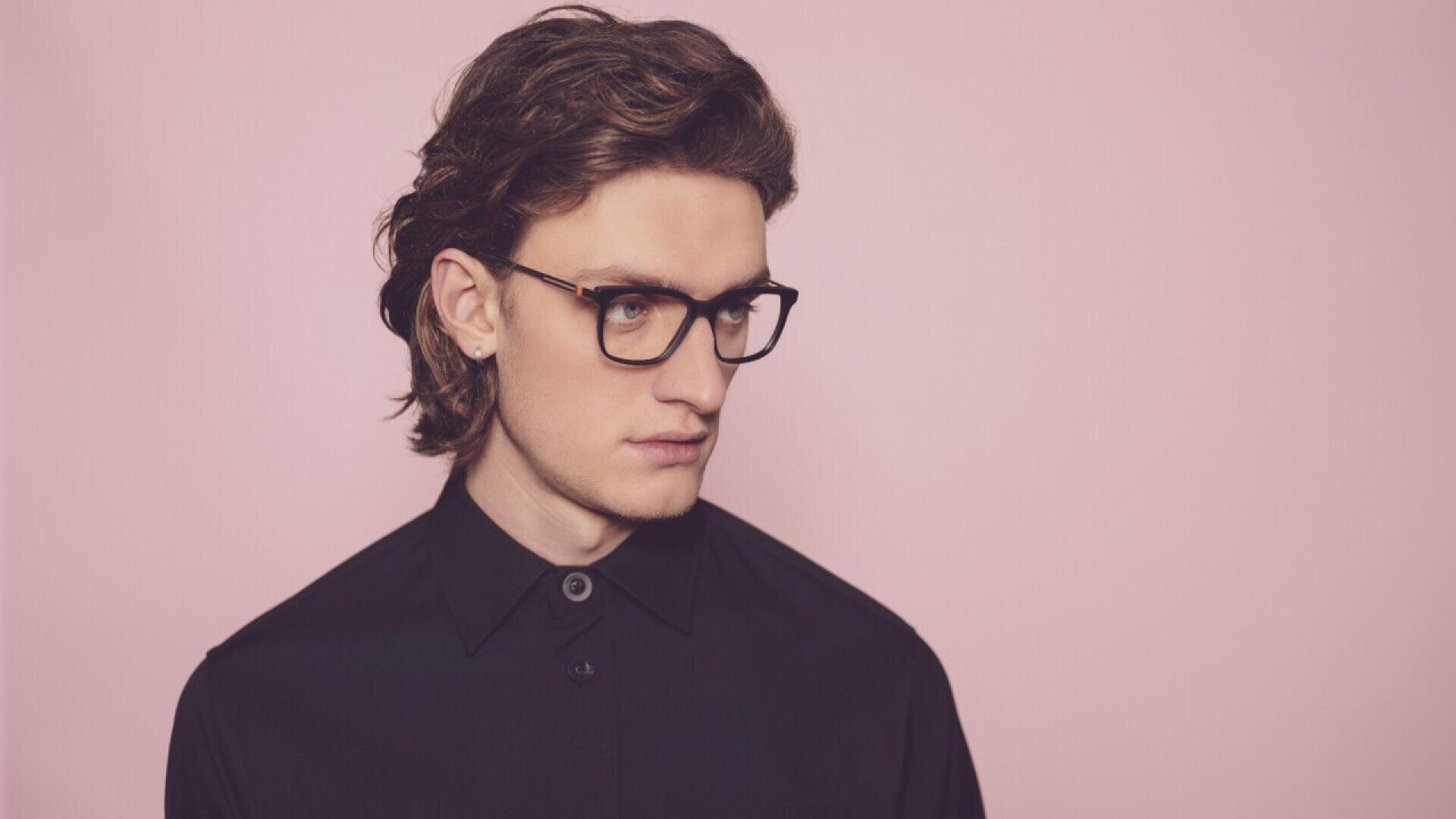A young person with wavy brown hair and glasses, wearing a black button-up shirt, stands against a plain light pink background, looking slightly to the side with a neutral expression.