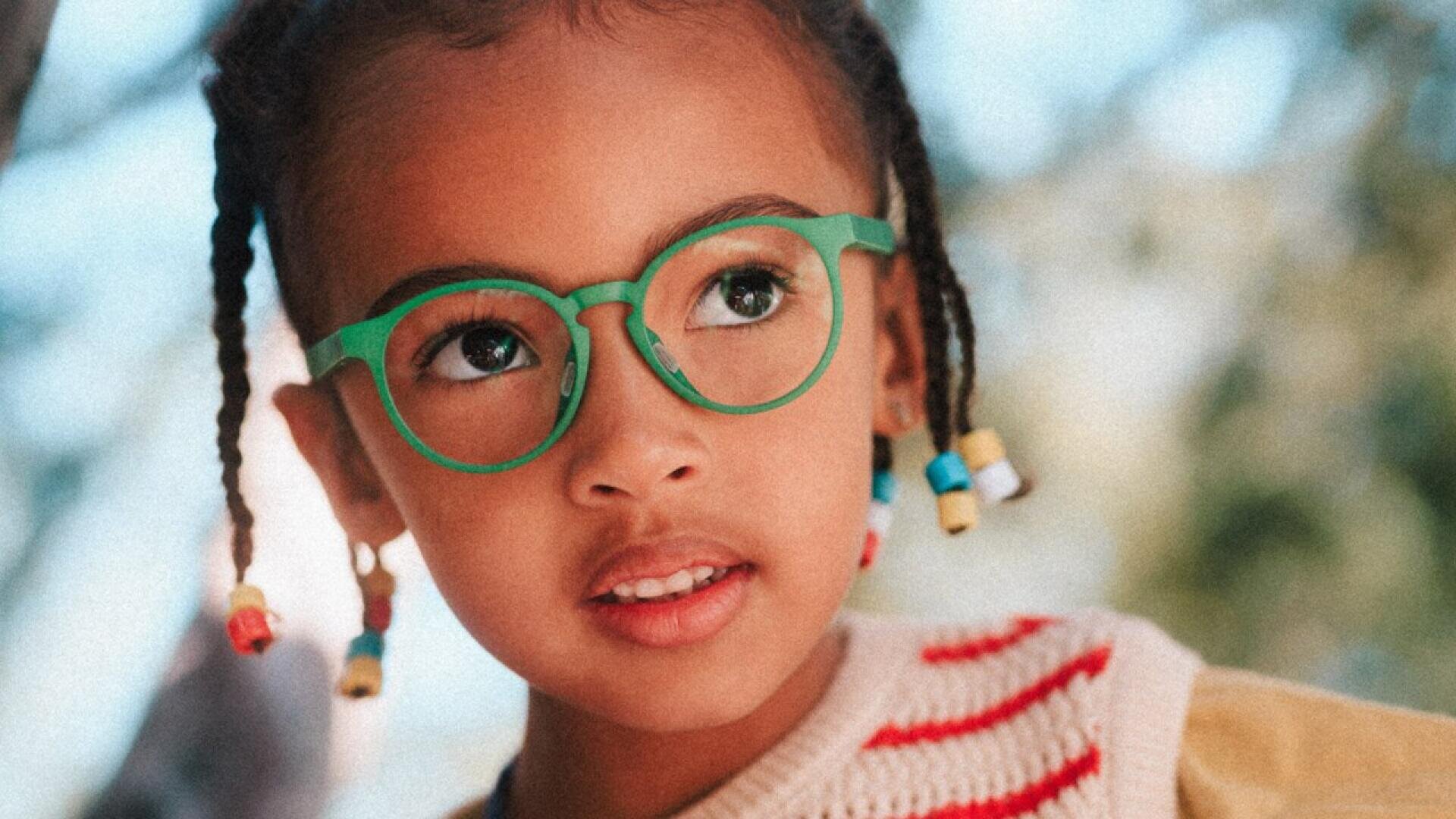 A young child with braided hair decorated with colorful beads wears green round glasses and a cream sweater with red stripes, looking slightly upwards with a curious expression.