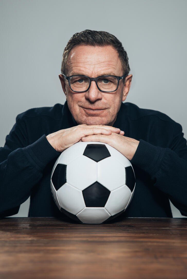 Ein Mann mittleren Alters mit Brille stützt seine Hände und sein Kinn auf einen klassischen schwarz-weißen Fußball und sitzt an einem Holztisch vor einem schlichten grauen Hintergrund.