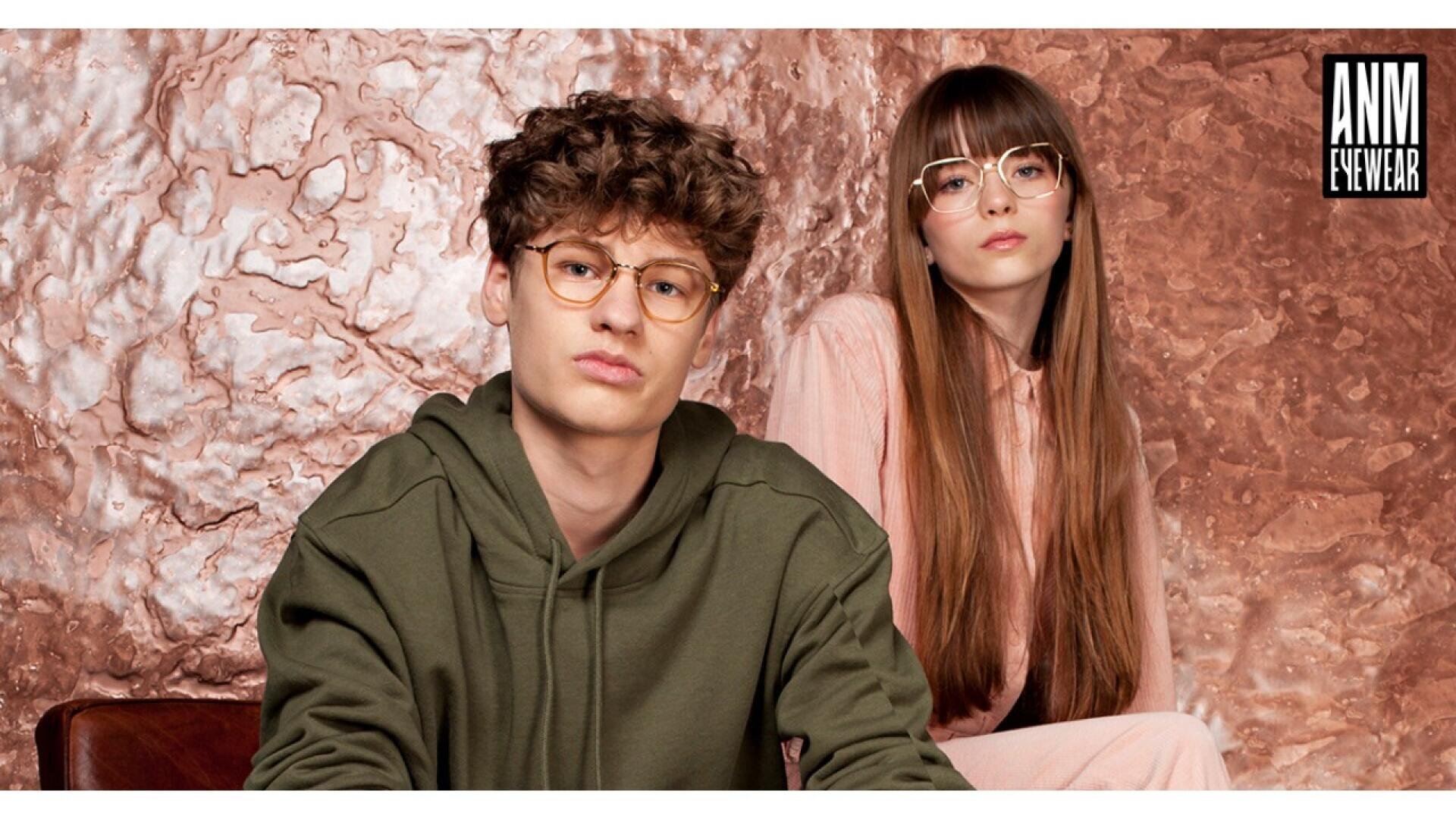 Two young adults wearing stylish eyeglasses pose in front of a textured copper background. The person on the left has curly hair and wears a green hoodie; the person on the right has long straight hair and wears pink clothing.