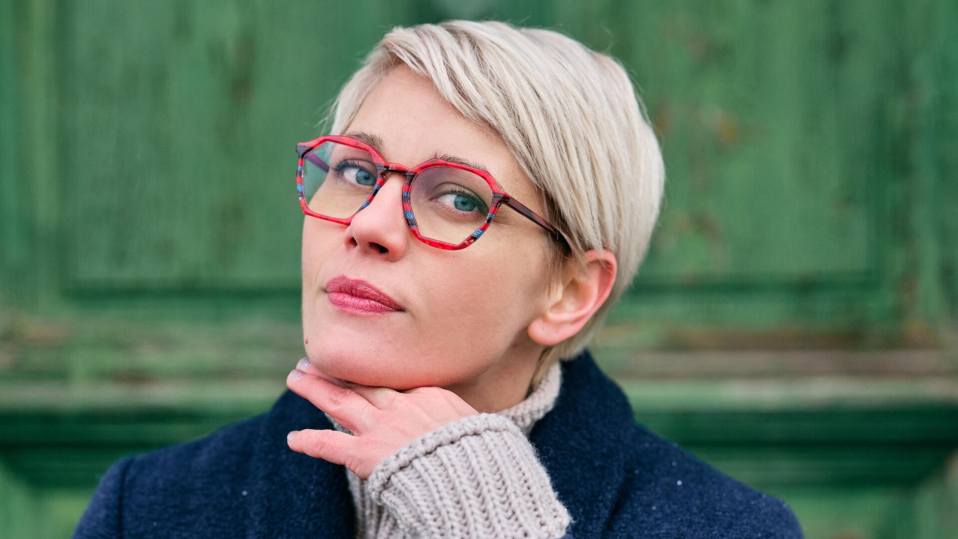 Eine Person mit kurzen blonden Haaren und roter Brille stützt ihr Kinn auf die Hand und blickt nachdenklich in die Kamera. Sie trägt einen marineblauen Mantel und einen grauen Pullover und steht vor einem grün strukturierten Hintergrund.