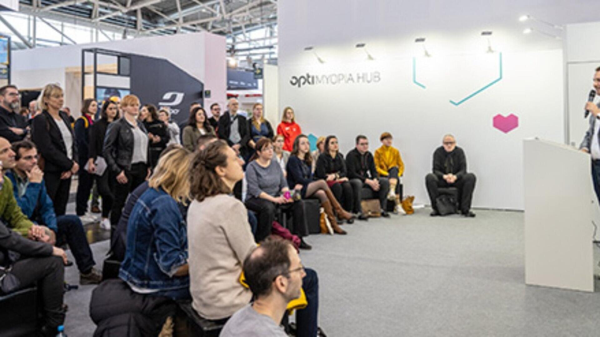 A group of people sits and stands attentively, listening to a speaker at a modern indoor event space labeled “OPTIMORA HUB.” The setting appears professional and collaborative.
