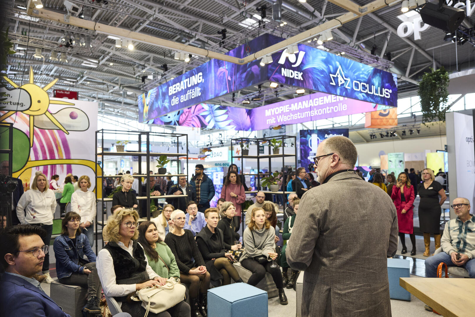 A man speaks to a seated audience at a brightly lit exhibition or trade show. Colorful banners for Nidek and Oculus hang above, and people listen attentively in a modern, busy indoor setting.