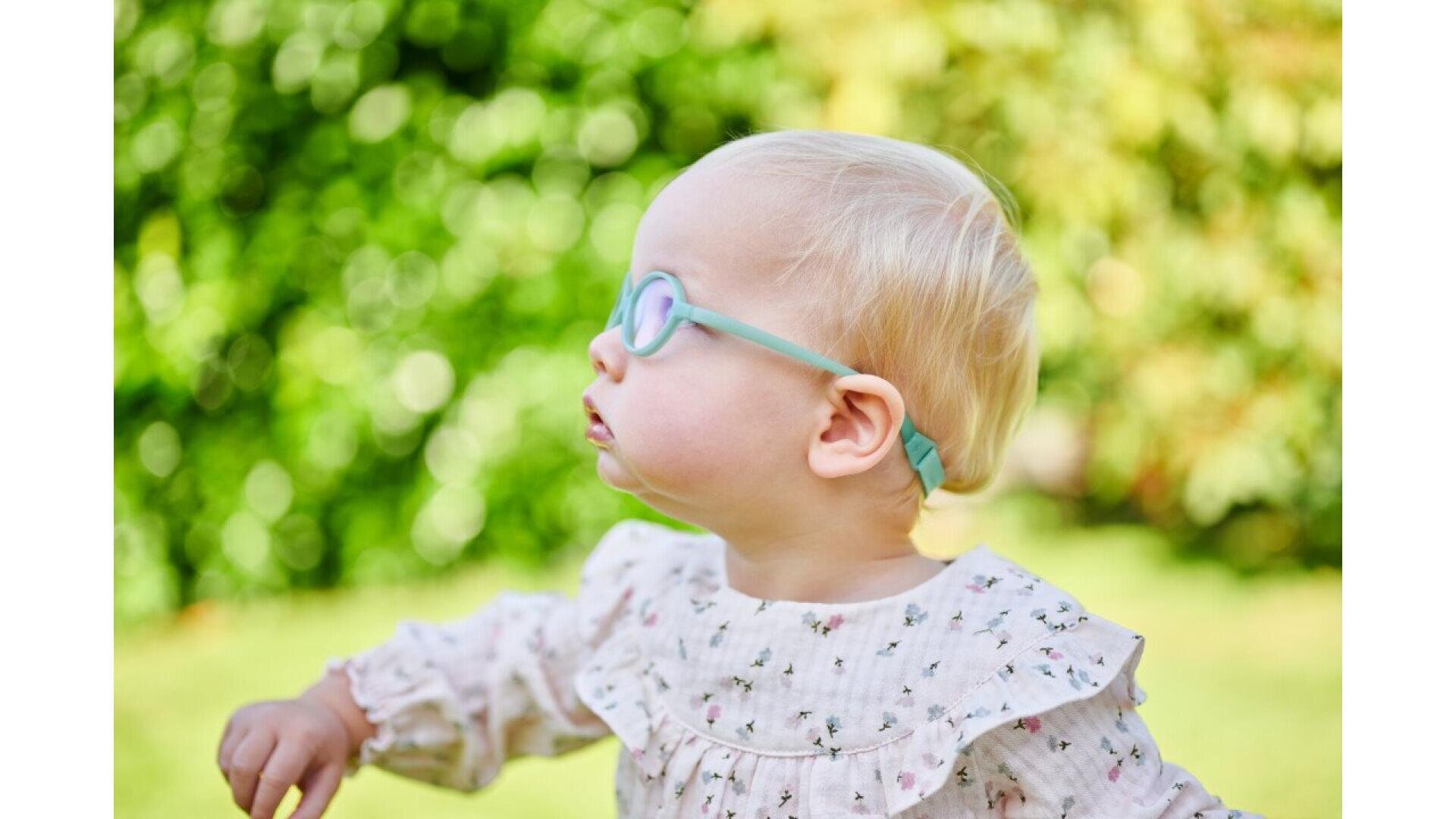 A young child with light blonde hair and blue glasses looks to the side while standing outdoors. The child wears a white floral top, and the background is filled with green foliage.