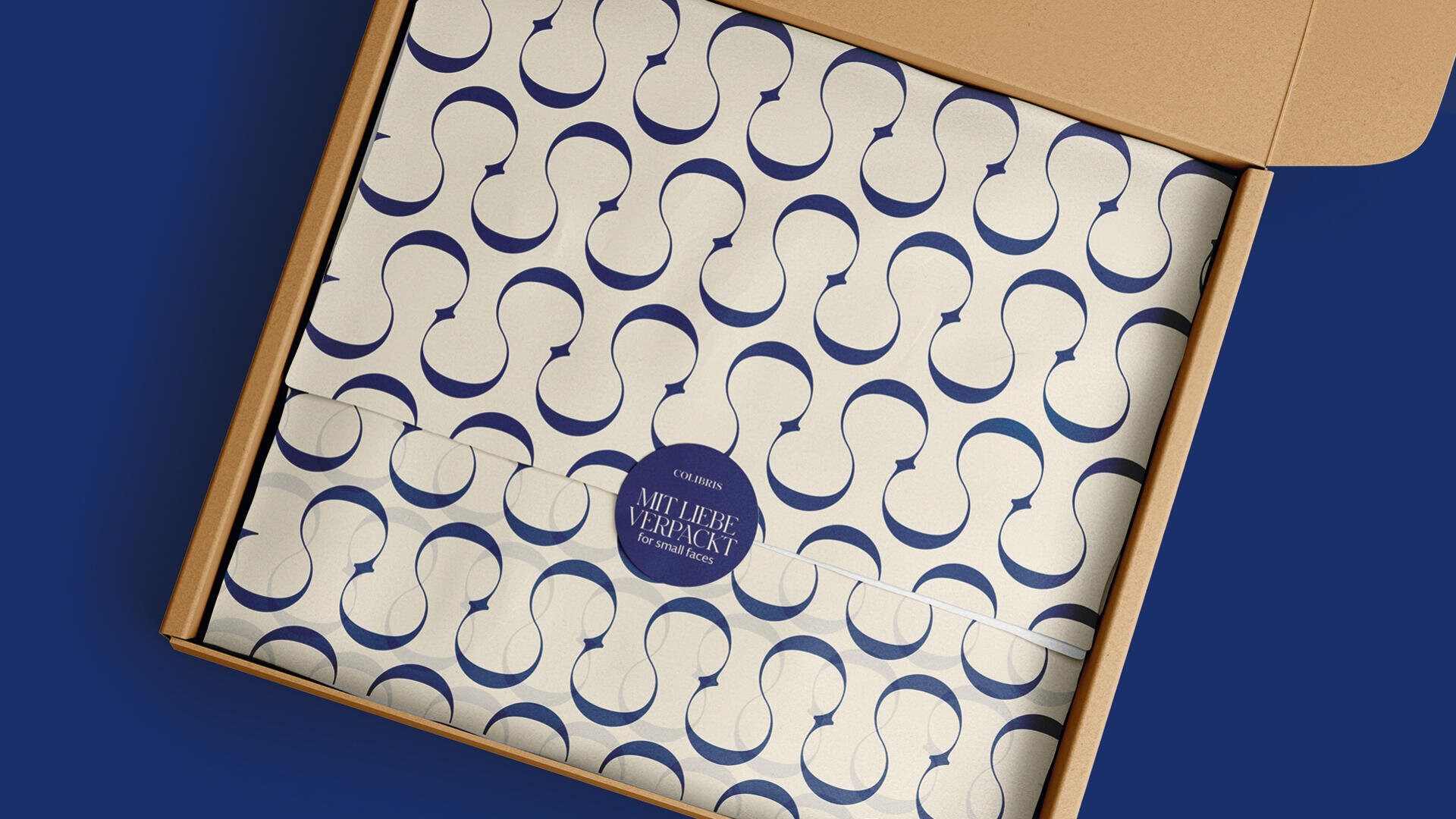 Ein Karton enthält ordentlich verpacktes Papier mit einem sich wiederholenden abstrakten blauen Muster und einem runden blauen Aufkleber mit der Aufschrift „MIT LIEBE VERPACKT“ in Weiß.