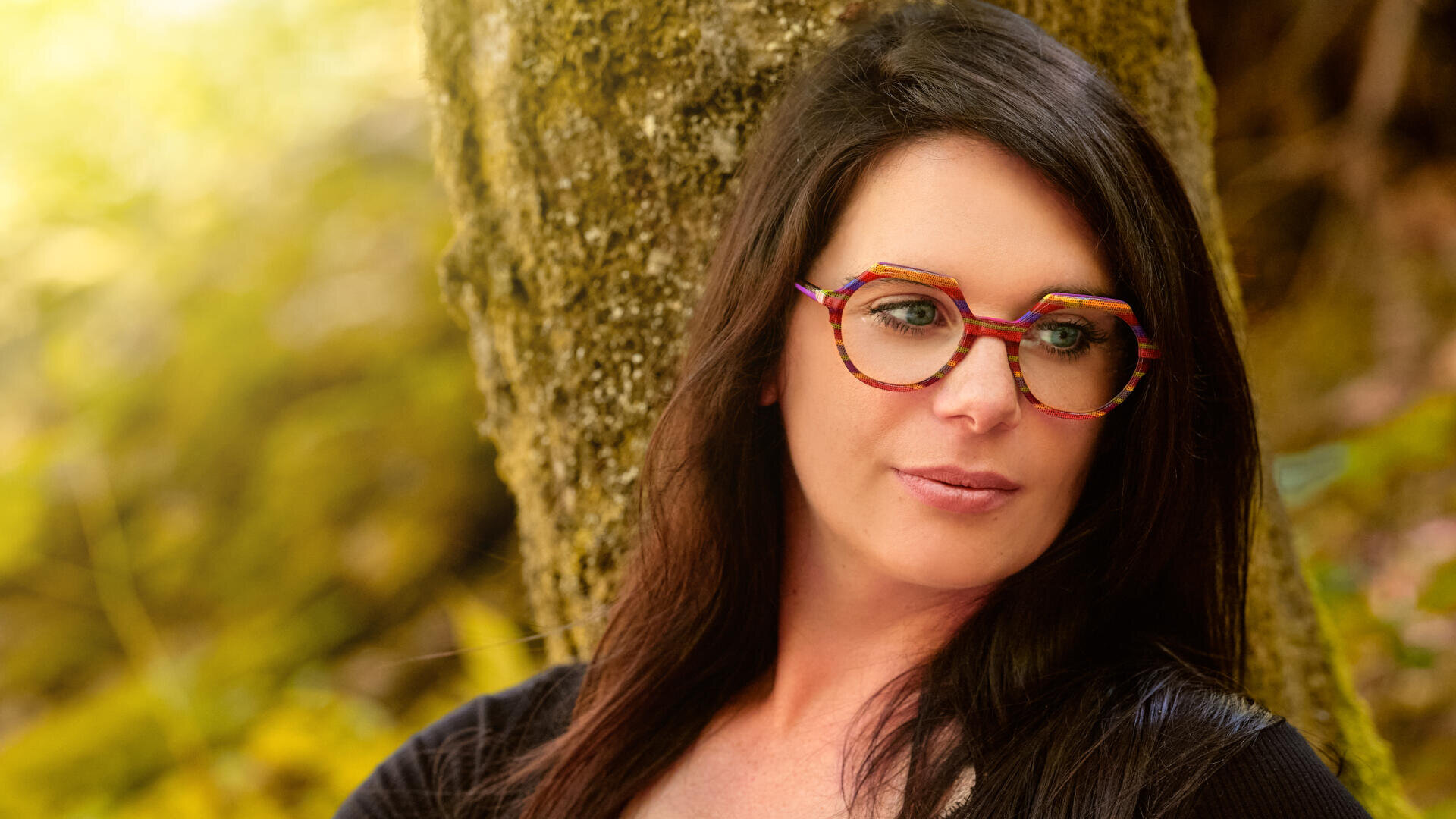 Eine Frau mit langem dunklem Haar und bunter, geometrischer Brille lehnt im Freien an einem Baum, umgeben von verschwommenem Grün und warmem Sonnenlicht.