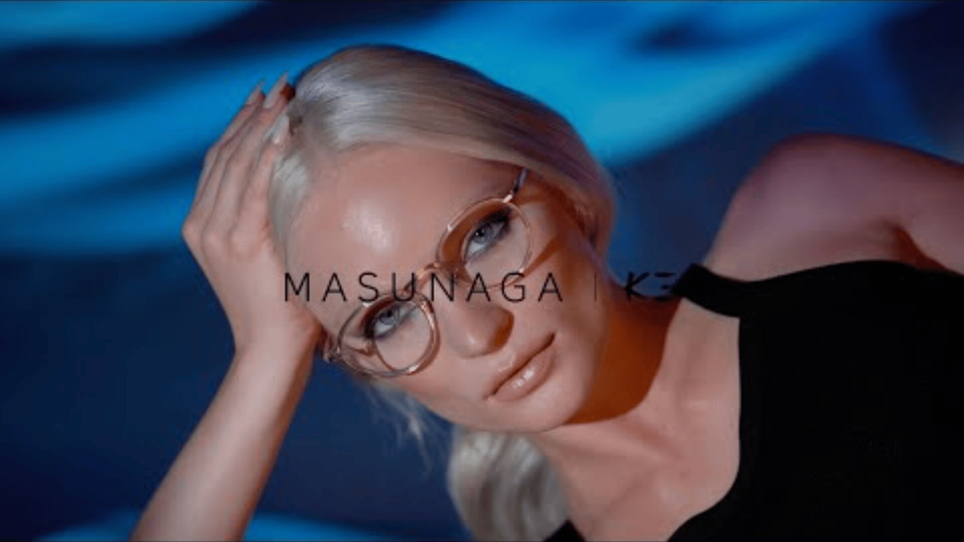 Eine Frau mit platinblondem Haar und Brille stützt ihren Kopf auf ihre Hand und blickt in die Kamera. Blaues Licht erzeugt einen dramatischen Hintergrund. Das Wort MASUNAGA ist über das Bild gelegt.