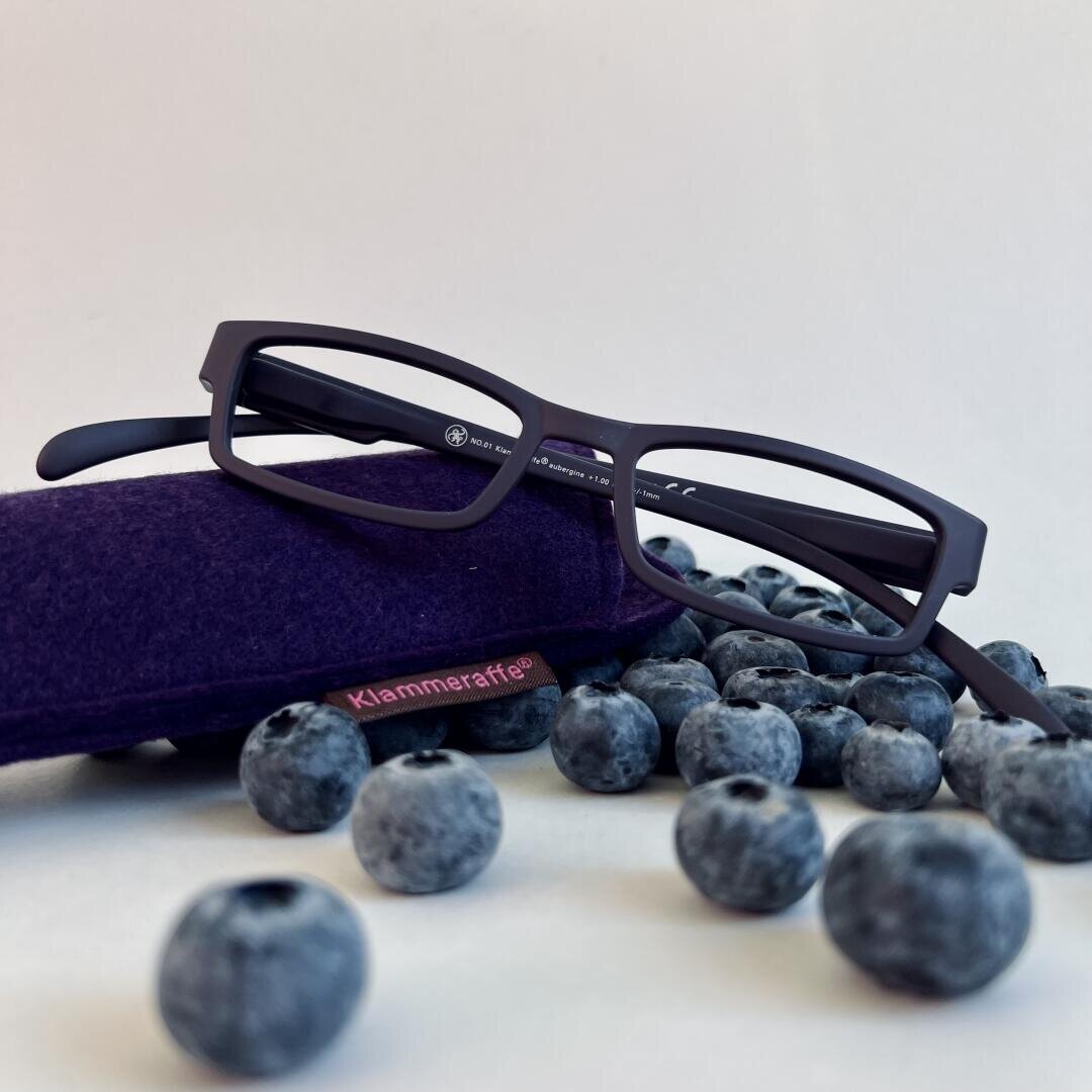 Eine schwarze rechteckige Brille liegt auf einem violetten Etui mit der Aufschrift „Klammeraffe“, umgeben von verstreuten frischen Blaubeeren auf einer weißen Oberfläche.