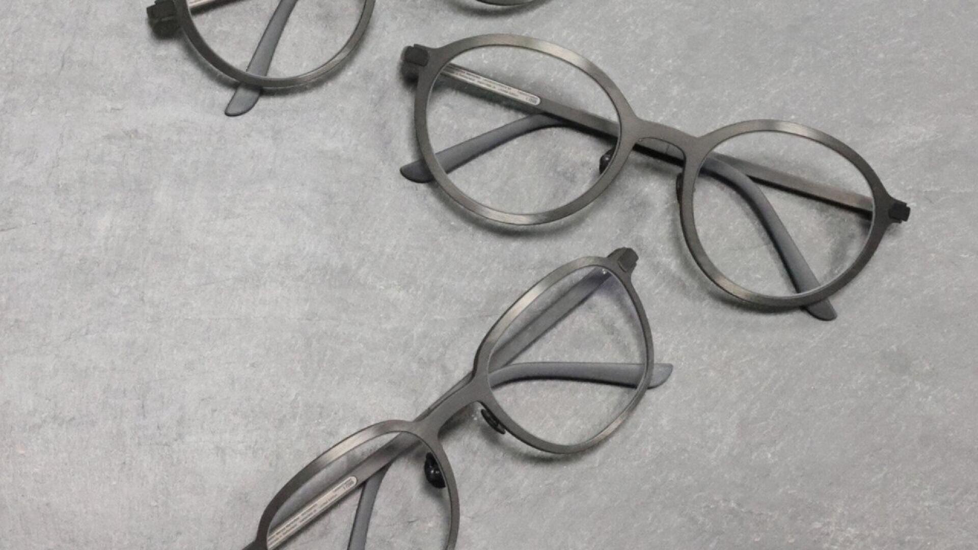 Three pairs of round, gray eyeglasses are placed on a light gray, textured surface. The glasses are arranged at different angles, with their arms open and lenses facing upward.