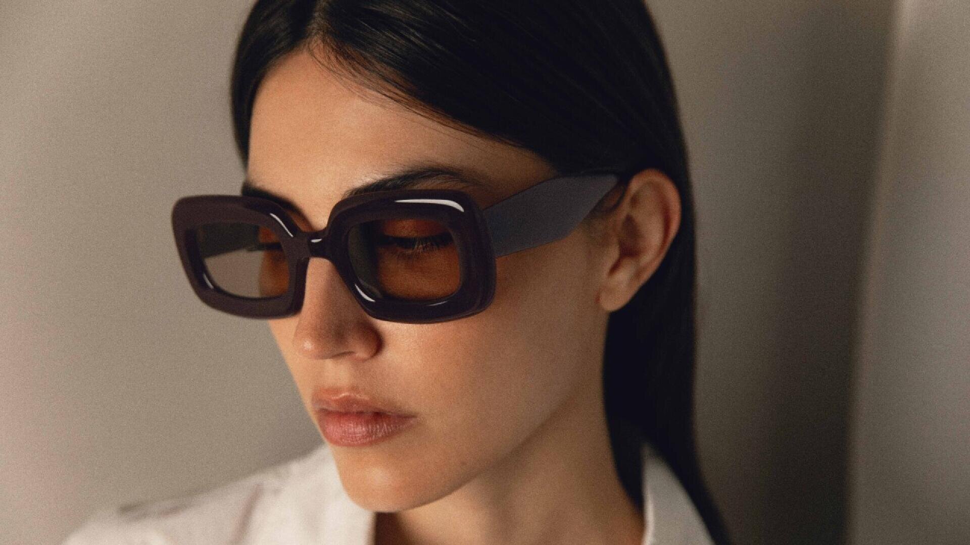 Eine Frau mit glattem, dunklem Haar trägt eine übergroße, eckige, dunkle Sonnenbrille und ein weißes Hemd mit Kragen und blickt in sanftem Licht nach unten und leicht zur Seite.