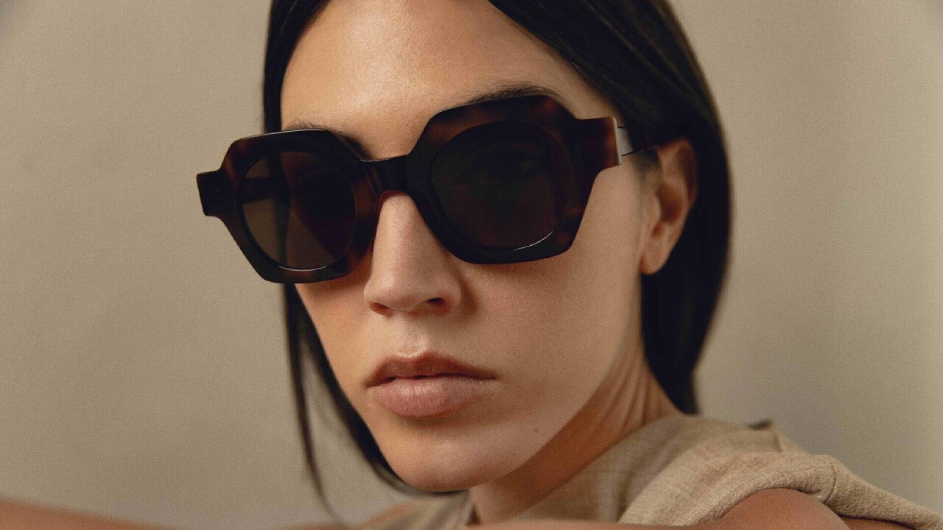 Eine Frau mit glattem, dunklem Haar trägt eine große, geometrische, dunkle Sonnenbrille und ein beiges ärmelloses Top. Sie blickt mit neutralem Gesichtsausdruck leicht an der Kamera vorbei. Der Hintergrund ist schlicht und sanft beleuchtet.