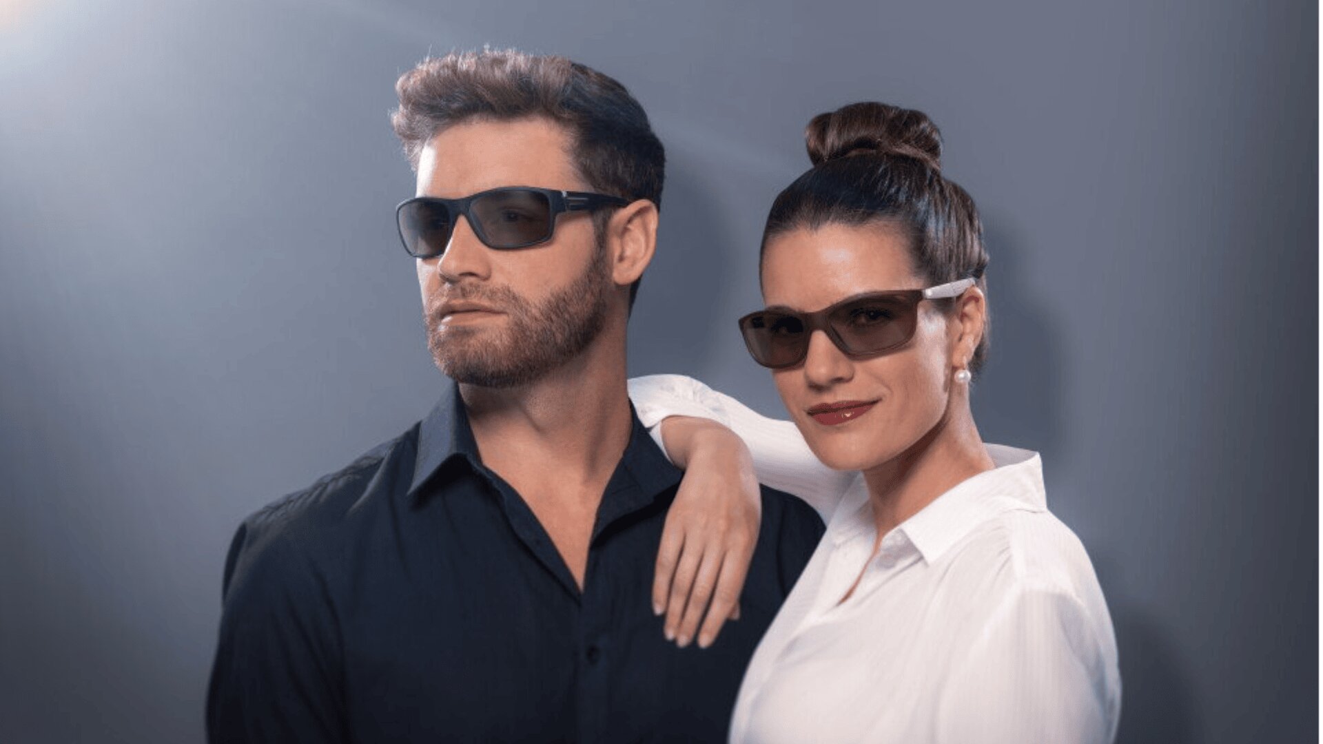 A man in a dark shirt and a woman in a white blouse, both wearing sunglasses, pose confidently against a gray gradient background. The woman has her arm resting on the mans shoulder.