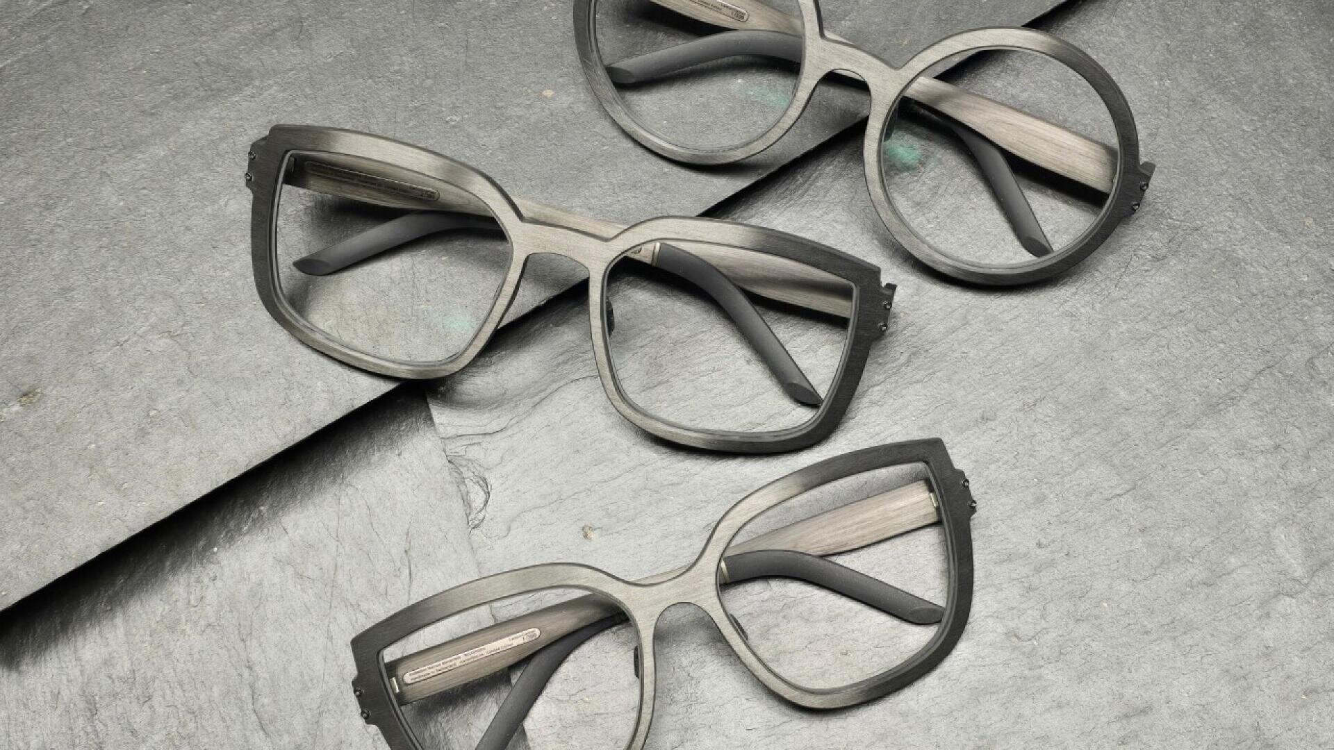 Three pairs of eyeglasses with thick, matte gray frames are arranged on a textured gray stone surface, positioned at different angles. The lenses are clear, and the glasses have wide black arms.