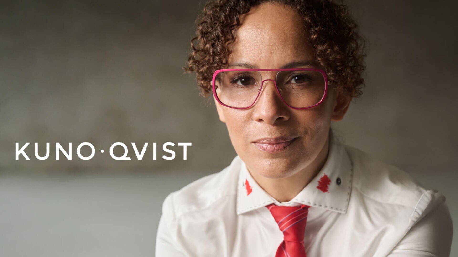 Eine Person mit lockigem Haar und großer rosa Brille trägt ein weißes Hemd mit roten Details und eine rote Krawatte. Der Hintergrund ist unscharf. Links neben der Person erscheint der Text KUNO QVIST.