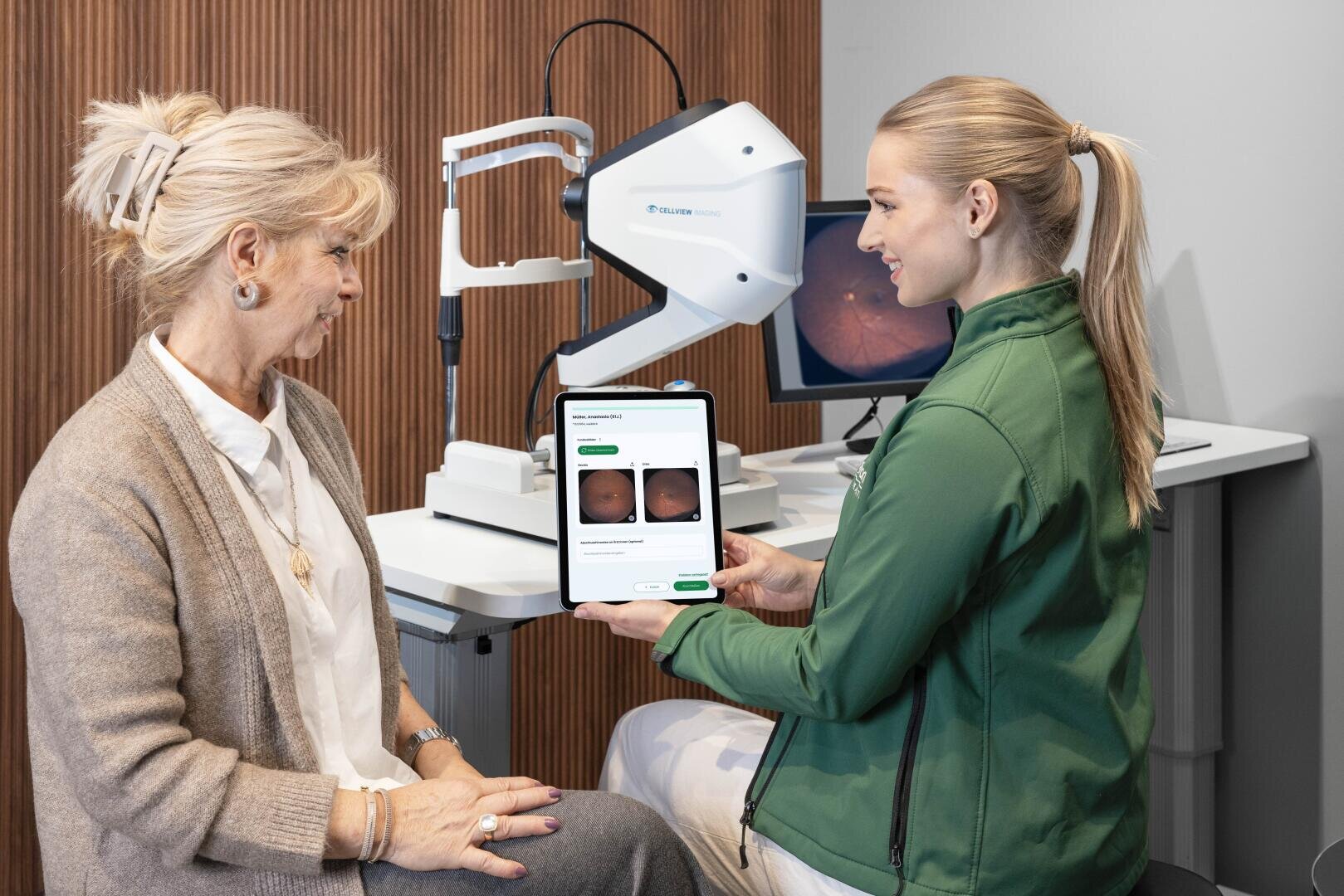 Eine medizinische Fachkraft zeigt einer älteren Frau ein Tablet mit Netzhautbildern und medizinischen Informationen während einer Augenuntersuchung in einer modernen Klinik mit Diagnosegeräten im Hintergrund.