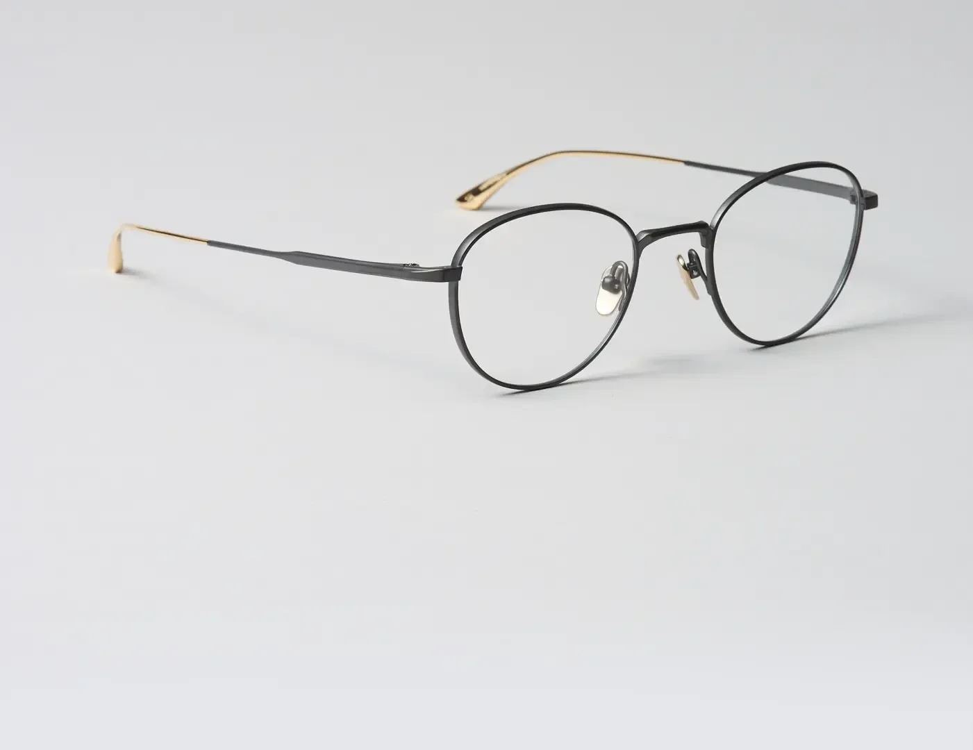 Eine Brille mit dünnem, schwarzem, rundem Rahmen und goldenen Bügeln, die auf einer schlichten, hellgrauen Oberfläche ruht.