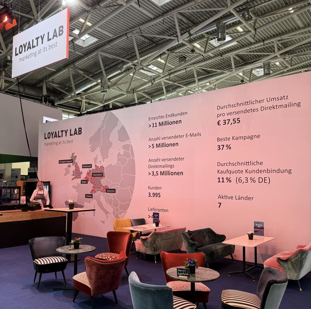 Ein Messestand von Loyalty Lab mit einer großen Wand, auf der Karten, Statistiken und Marketingkennzahlen auf Deutsch abgebildet sind. Es gibt rosafarbene Stühle, Tische und einen Tresen mit einer Mitarbeiterin. Der Stand ist gut beleuchtet und modern.