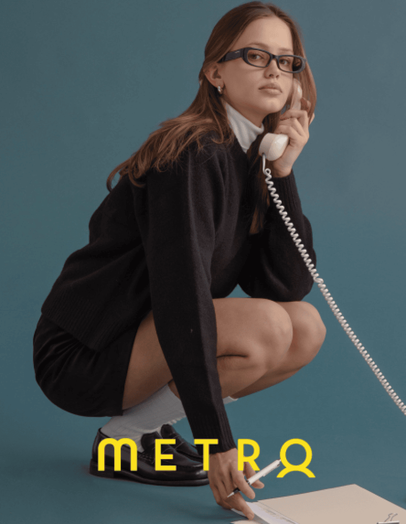 Eine Frau mit Brille, schwarzem Pullover und weißem Rollkragenpullover geht in die Hocke und hält ein schnurgebundenes Telefon an ihr Ohr. Sie blickt nach vorne, zu ihren Füßen liegen Dokumente und das Wort METRO in fetten gelben Buchstaben.