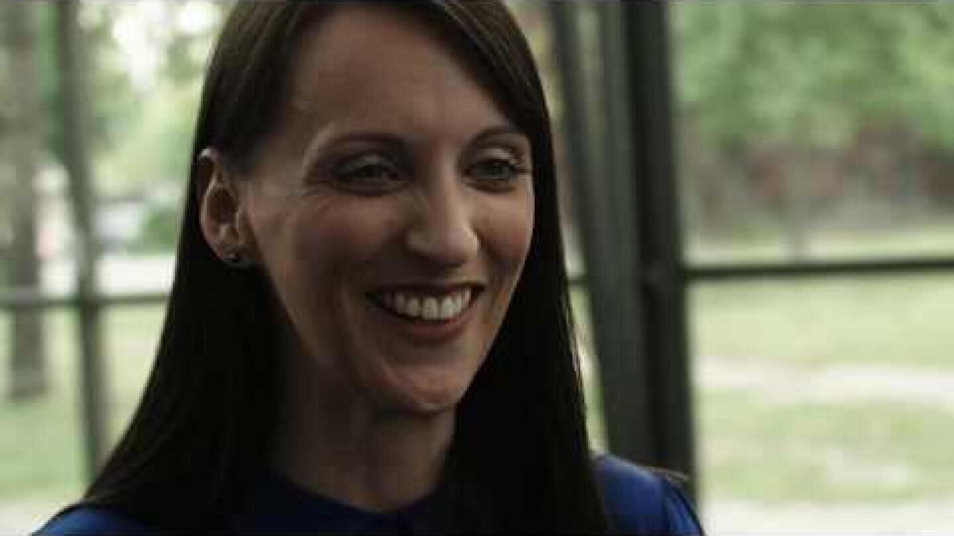 A woman with long dark hair smiles warmly in an interior in which large windows and green areas can be seen in the blurred background.