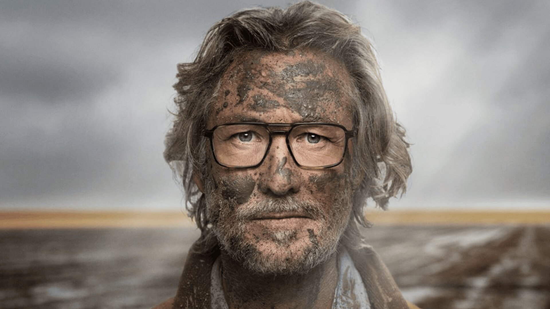 Ein Mann mit grauem Haar, Brille und Bart steht im Freien mit Schlammspritzern im Gesicht. Der Hintergrund ist eine verschwommene, karge Landschaft unter einem bewölkten Himmel.
