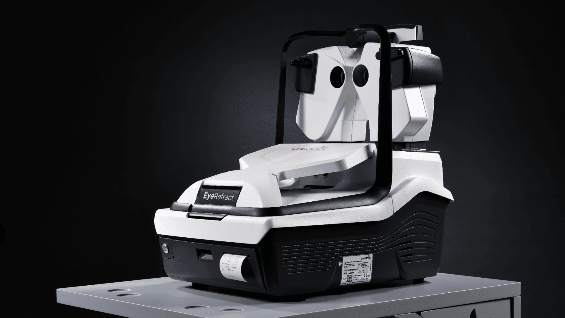 Ein modernes augenärztliches Diagnosegerät, hauptsächlich weiß mit schwarzen Akzenten, wird auf einen Metalltisch gestellt. Das Gerät ist mit einer Kinnstütze und Linsen für Augenuntersuchungen ausgestattet, die für Sehtests oder Netzhautuntersuchungen verwendet werden.