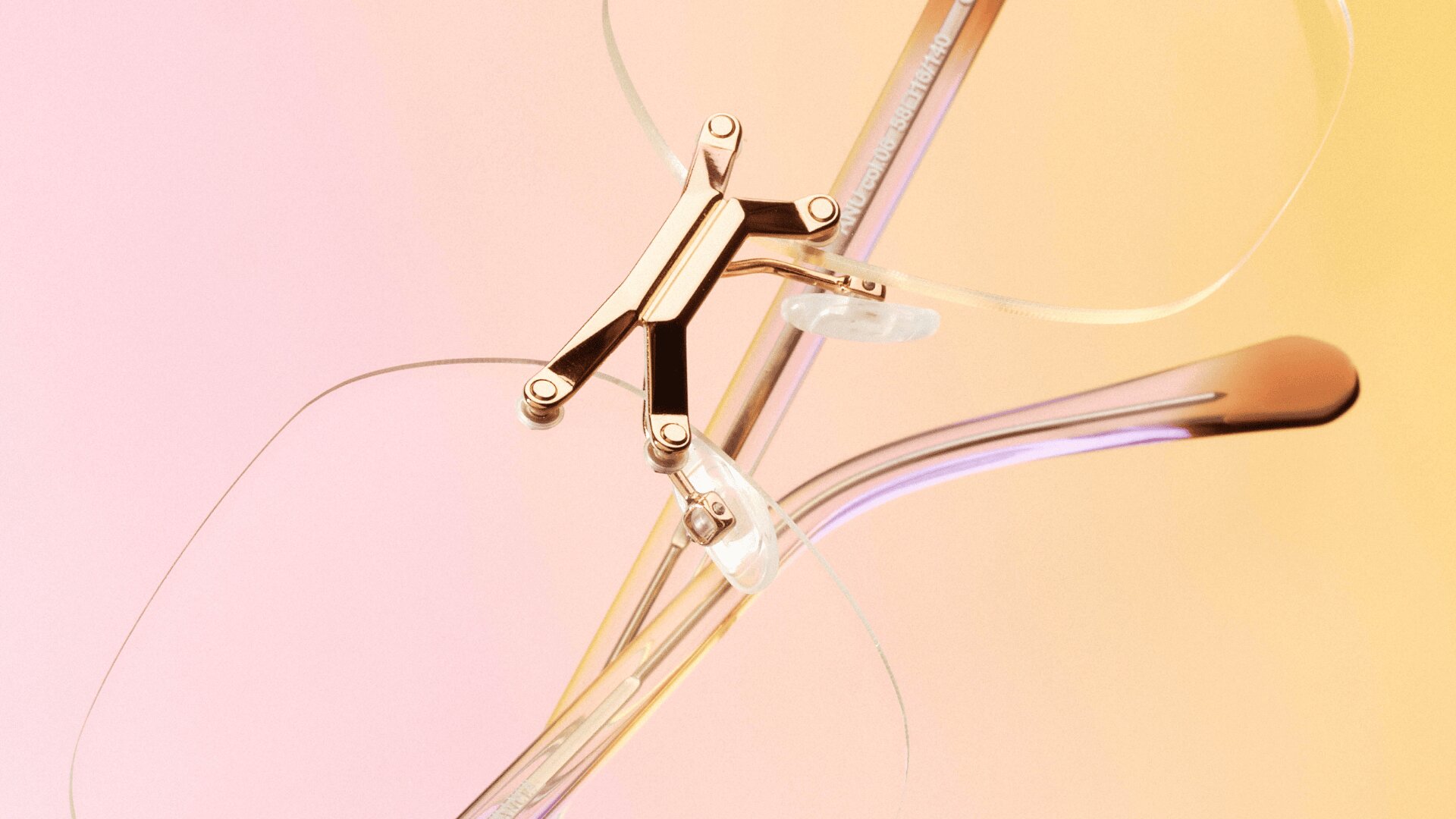 Nahaufnahme einer randlosen Brille mit goldenen Metallbügeln und klaren Nasenpads vor einem Hintergrund mit sanftem rosa und gelbem Farbverlauf.