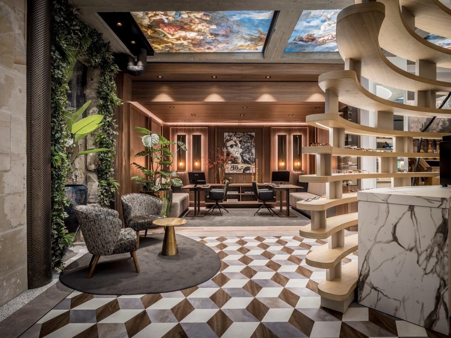 Moderne, stilvolle Hotellobby mit geometrischen Bodenfliesen, Marmorrezeption, Holzspindeltreppe, üppigen Grünpflanzen und einer gemütlichen Sitzecke. An der Decke befindet sich ein künstlerisches Wandgemälde; warme Beleuchtung schafft eine einladende Atmosphäre.
