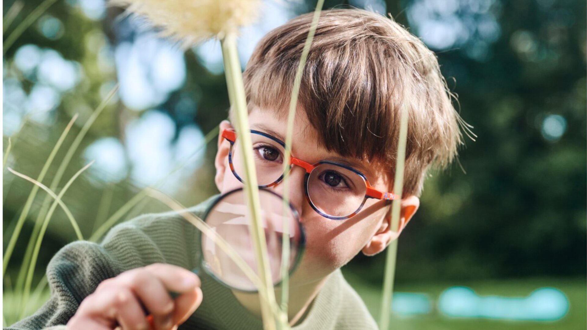 A young boy with brown hair and glasses examines a dandelion up close with a magnifying glass in an outdoor, grassy area.